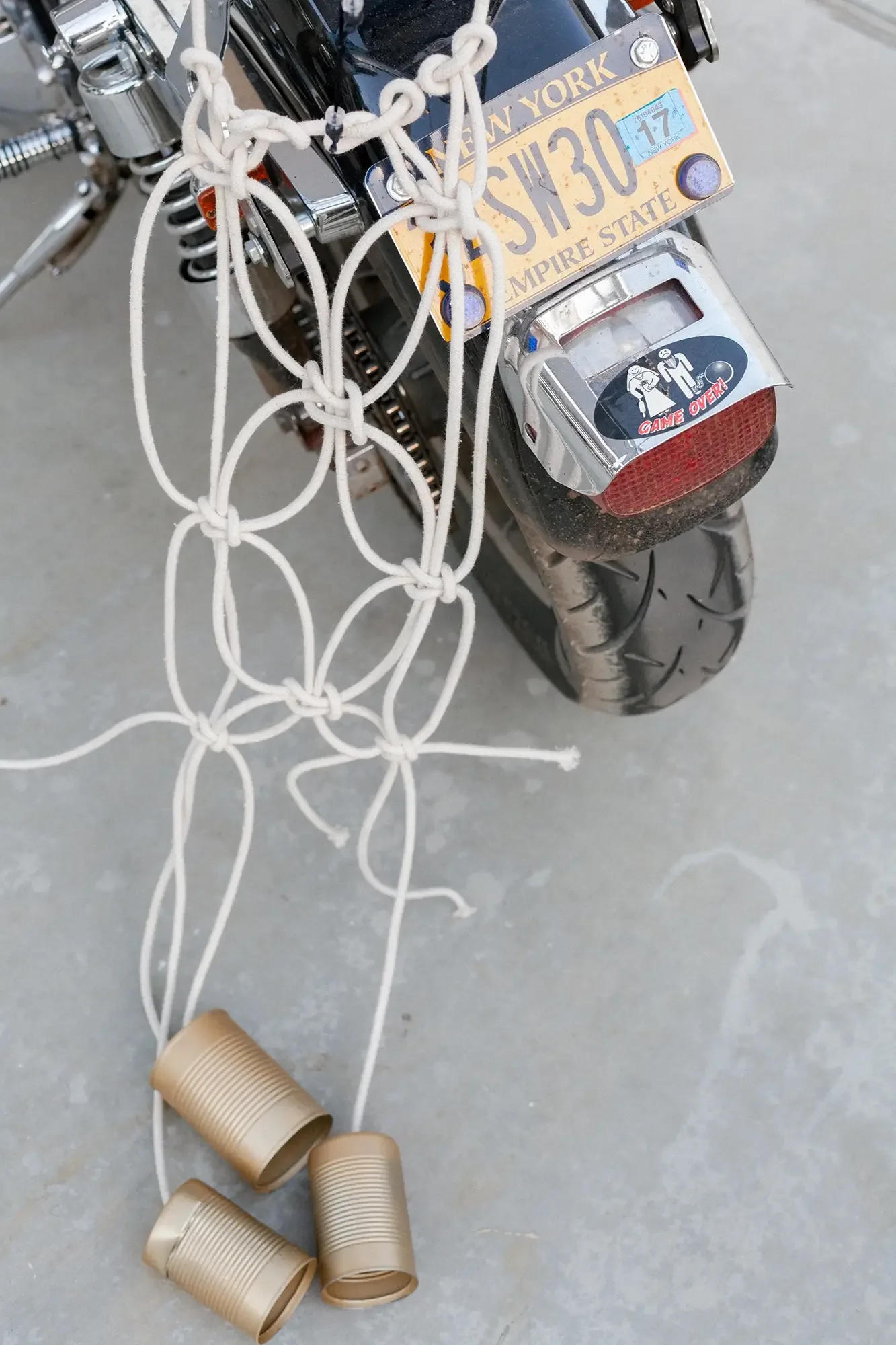 A motorcycle with a tied net covering three empty cans, a yellow New York license plate, and a 'Game Over' sticker on the rear.