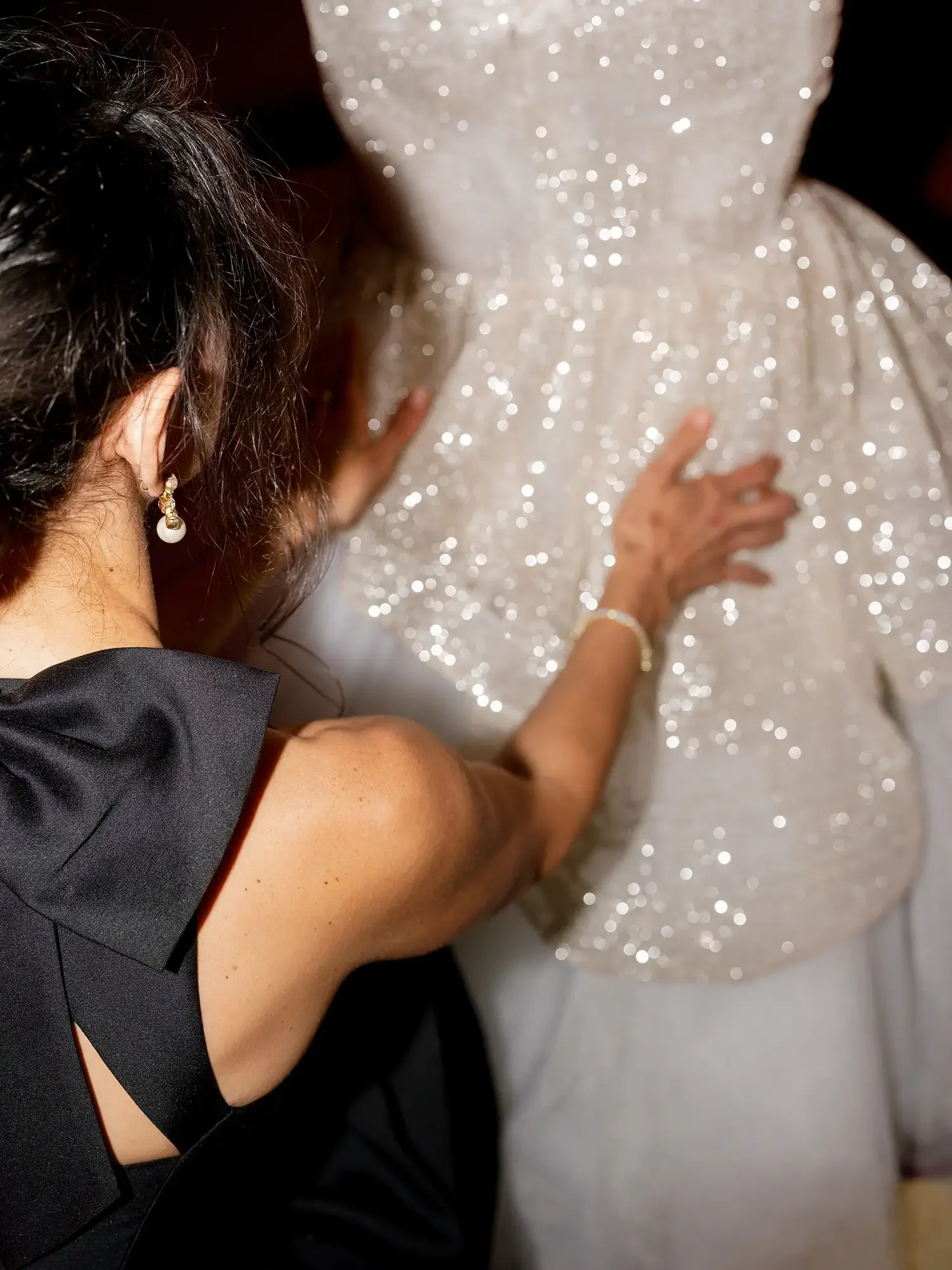 A woman with dark hair and pearl earrings is adjusting a glittery, sequined wedding dress or evening gown.