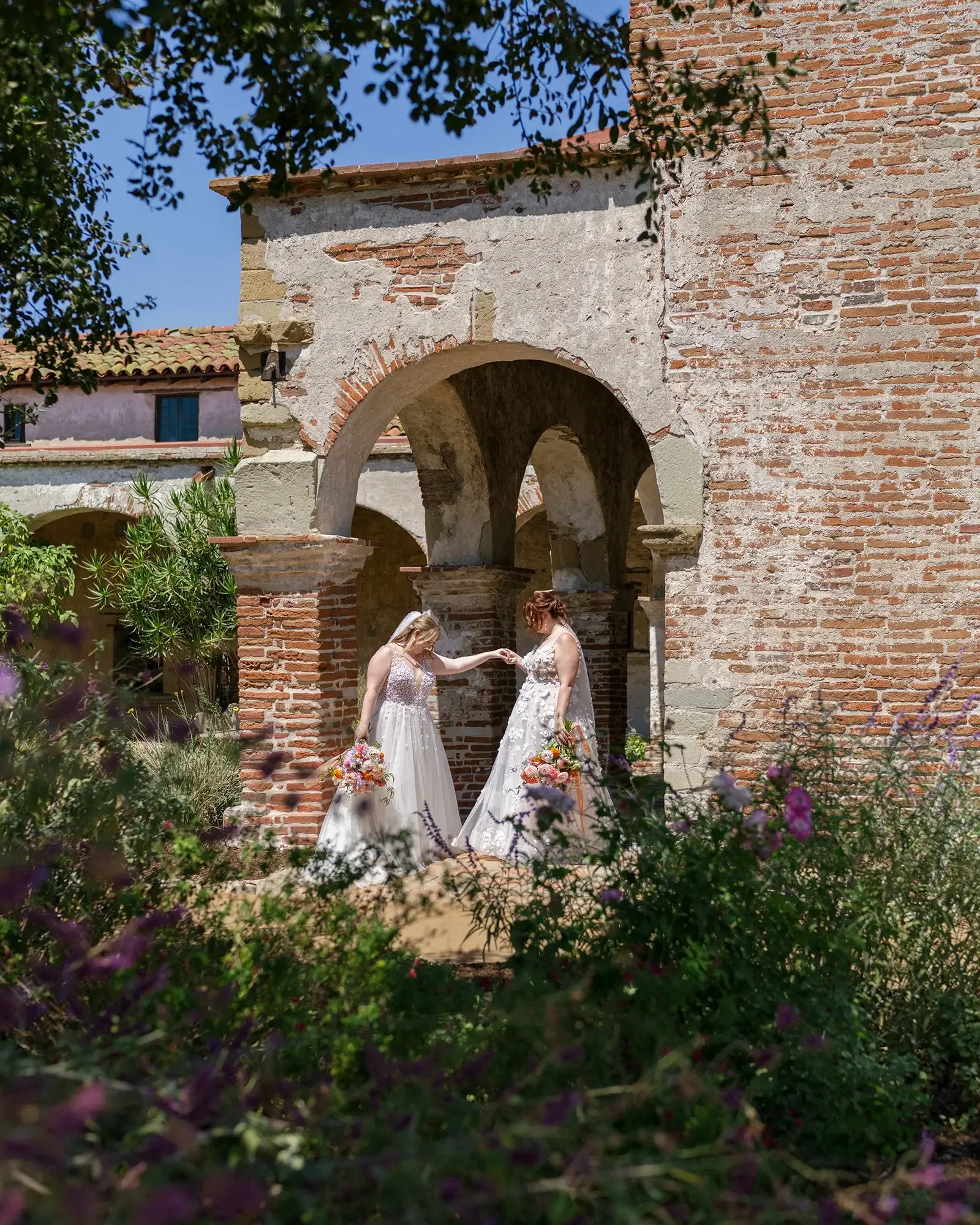 Two brides in white wedding gowns holding bouquets stand under a brick archway surrounded by greenery, with an old stone building and blue sky in the background.