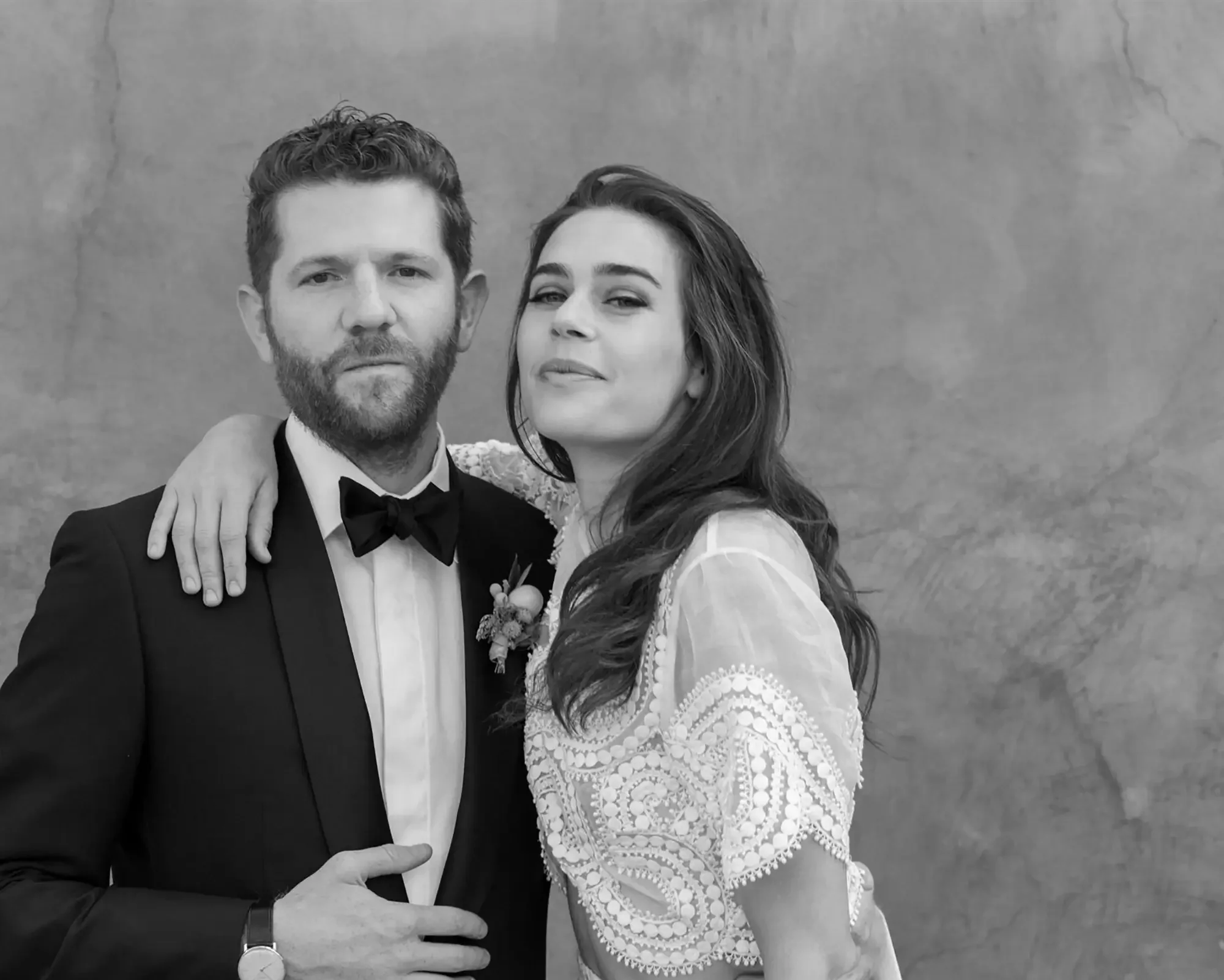 A black and white photo of a bride and groom posing together. The man is dressed in a tuxedo with a bow tie, and the woman is wearing a lace dress. They are standing against a plain background.