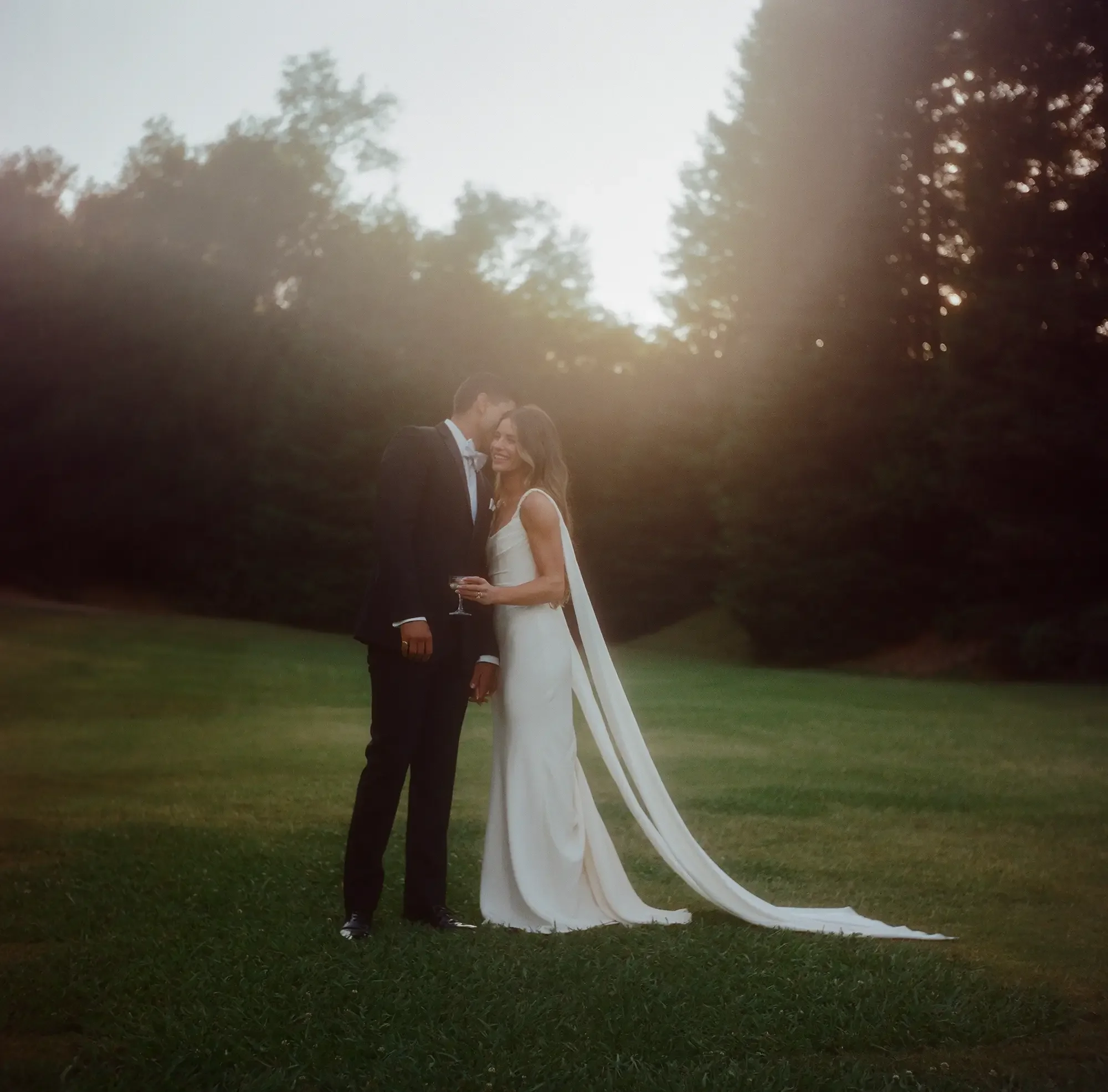 A bride and groom in wedding attire sharing a moment outdoors during sunset, with trees in the background.