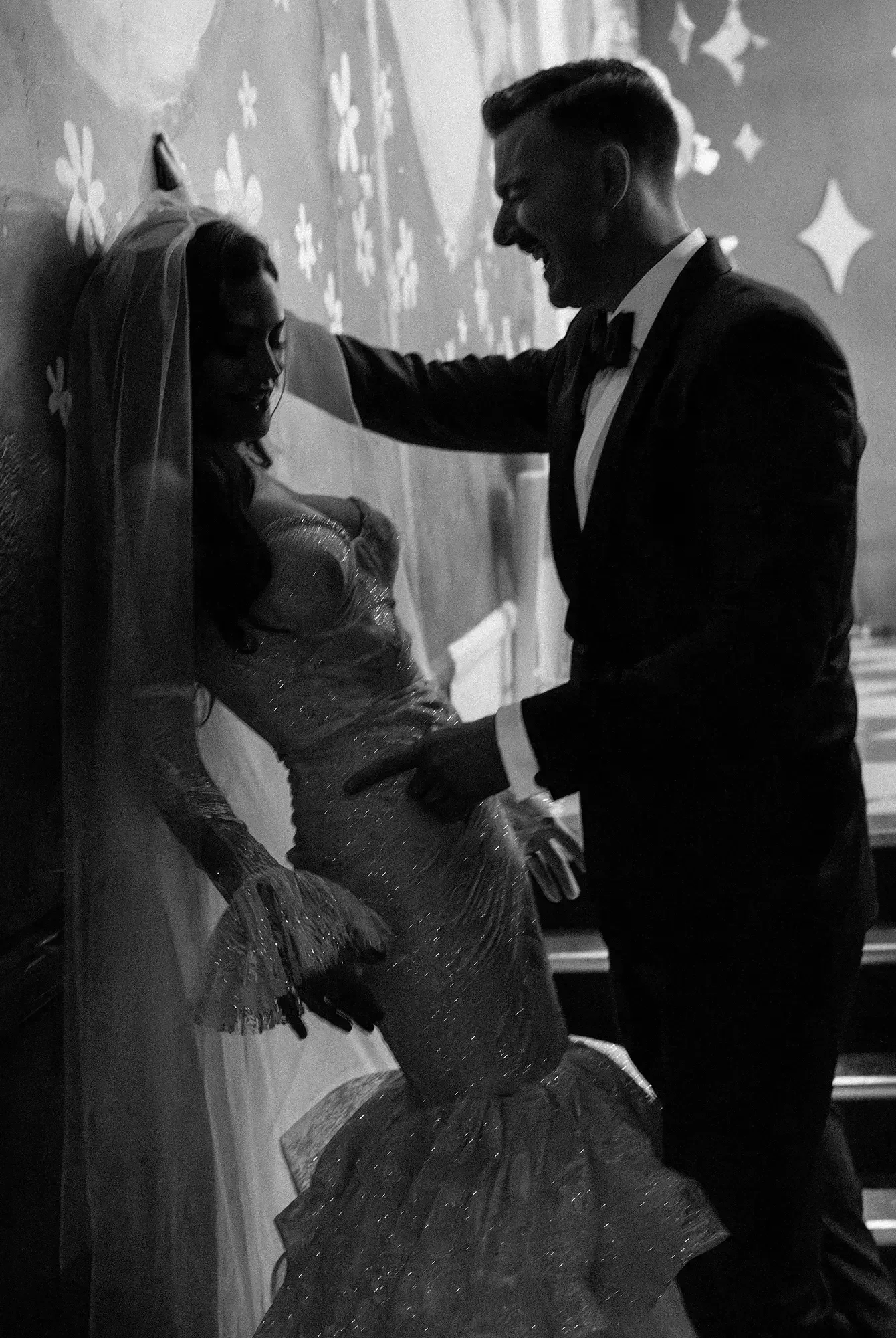 A black and white photo of a smiling groom in a tuxedo dancing with his bride who is wearing a sparkly dress and veil, leaning against a wall decorated with stars and flowers.