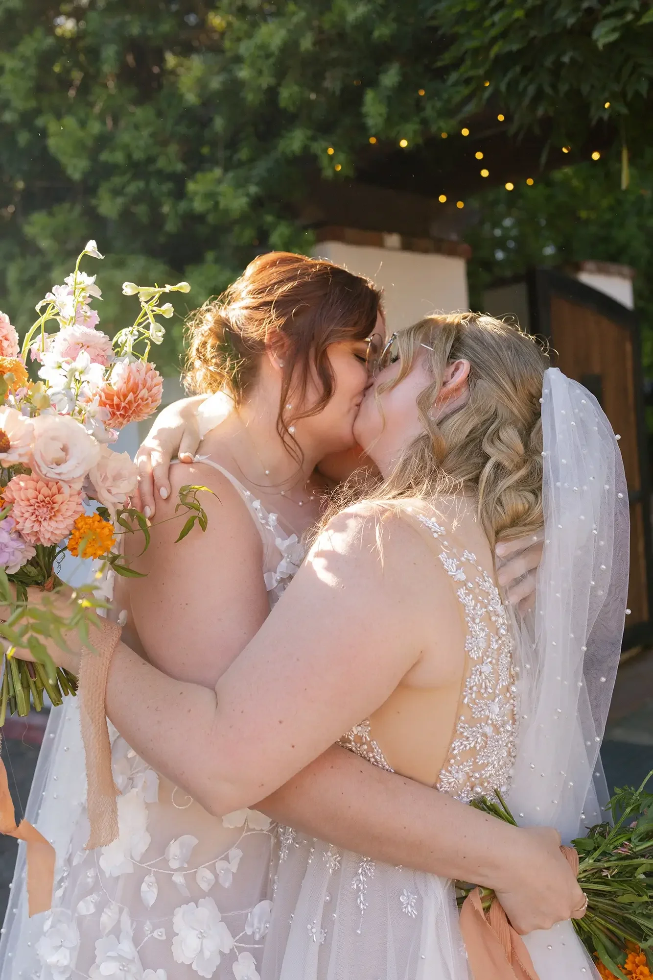 Two brides in wedding dresses sharing a kiss outdoors, one holding a bouquet of flowers, with sunlight filtering through trees and small string lights in the background.