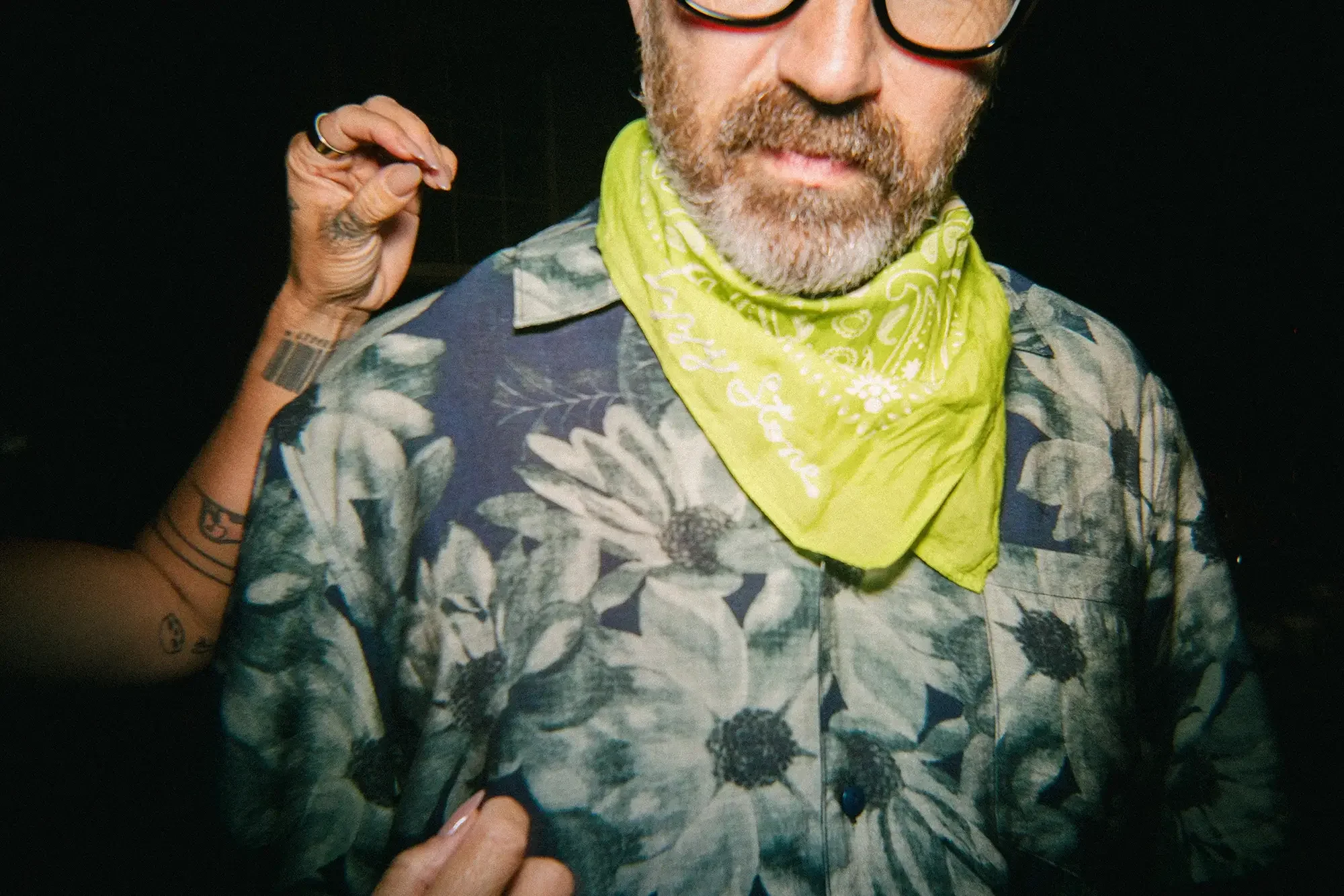Close-up of a man wearing a floral shirt, yellow bandana, and glasses, with part of another person's arm and hand in the background.