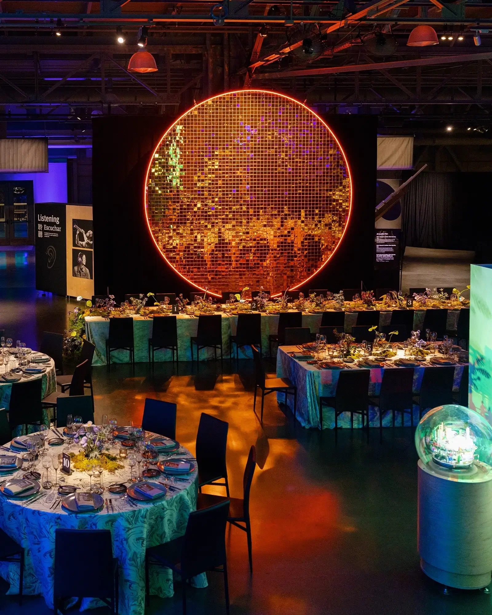 A dark wedding reception decorated with round tables covered in colorful tablecloths, set with plates, glassware, and floral centerpieces. 