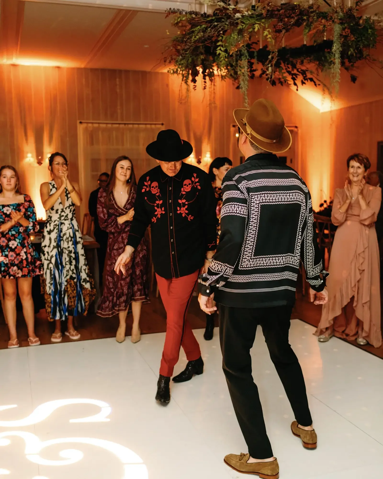 Two grooms dancing at their wedding in a warmly lit room with wooden walls and hanging greenery.