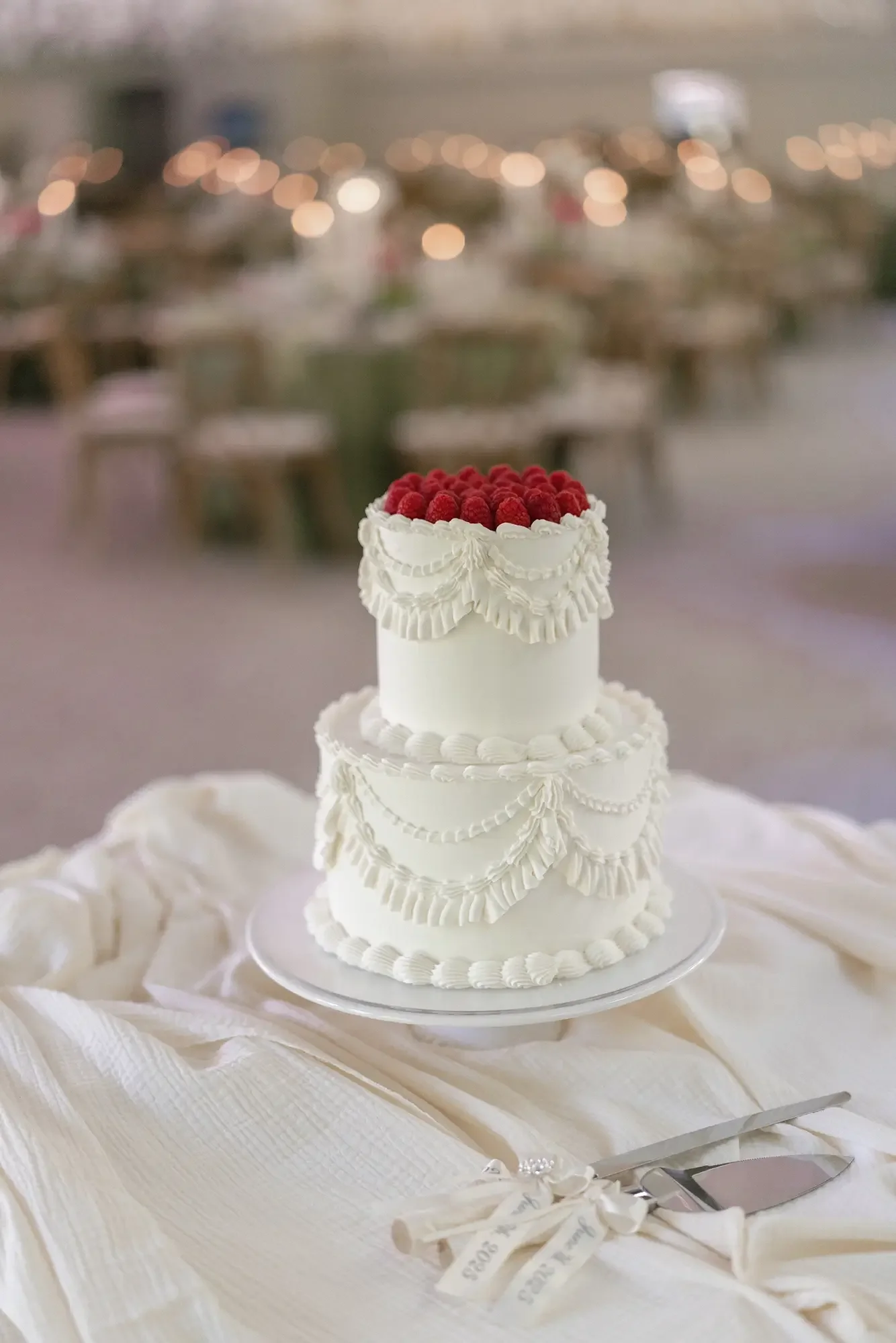 A white, two-tiered wedding cake decorated with piped icing and topped with raspberries, set on a white tablecloth with cake cutting tools nearby, in a blurred event hall background.