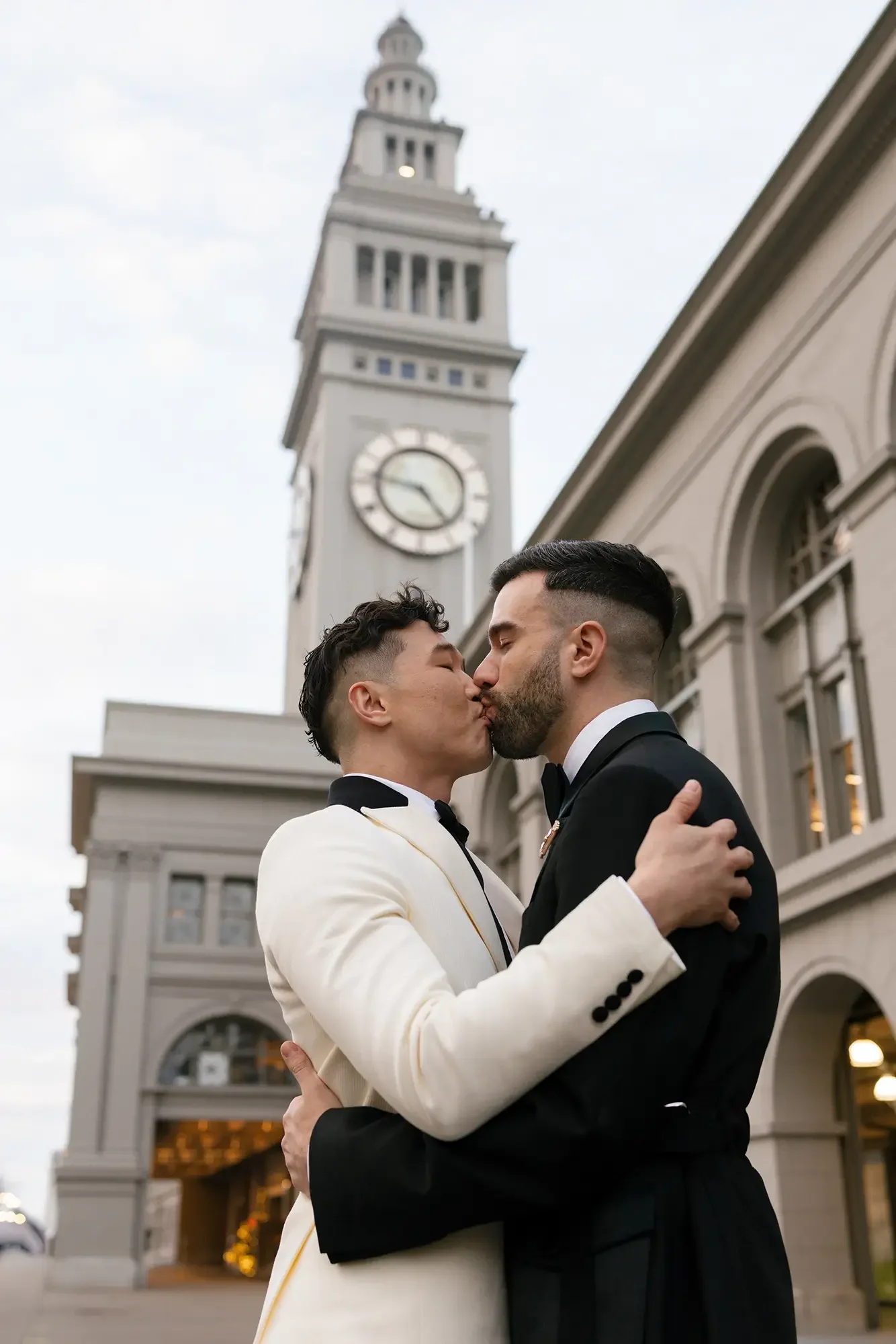 Two grooms kissing in front of a clock tower in San Francisco with a classic building behind them, both dressed in tuxedos.