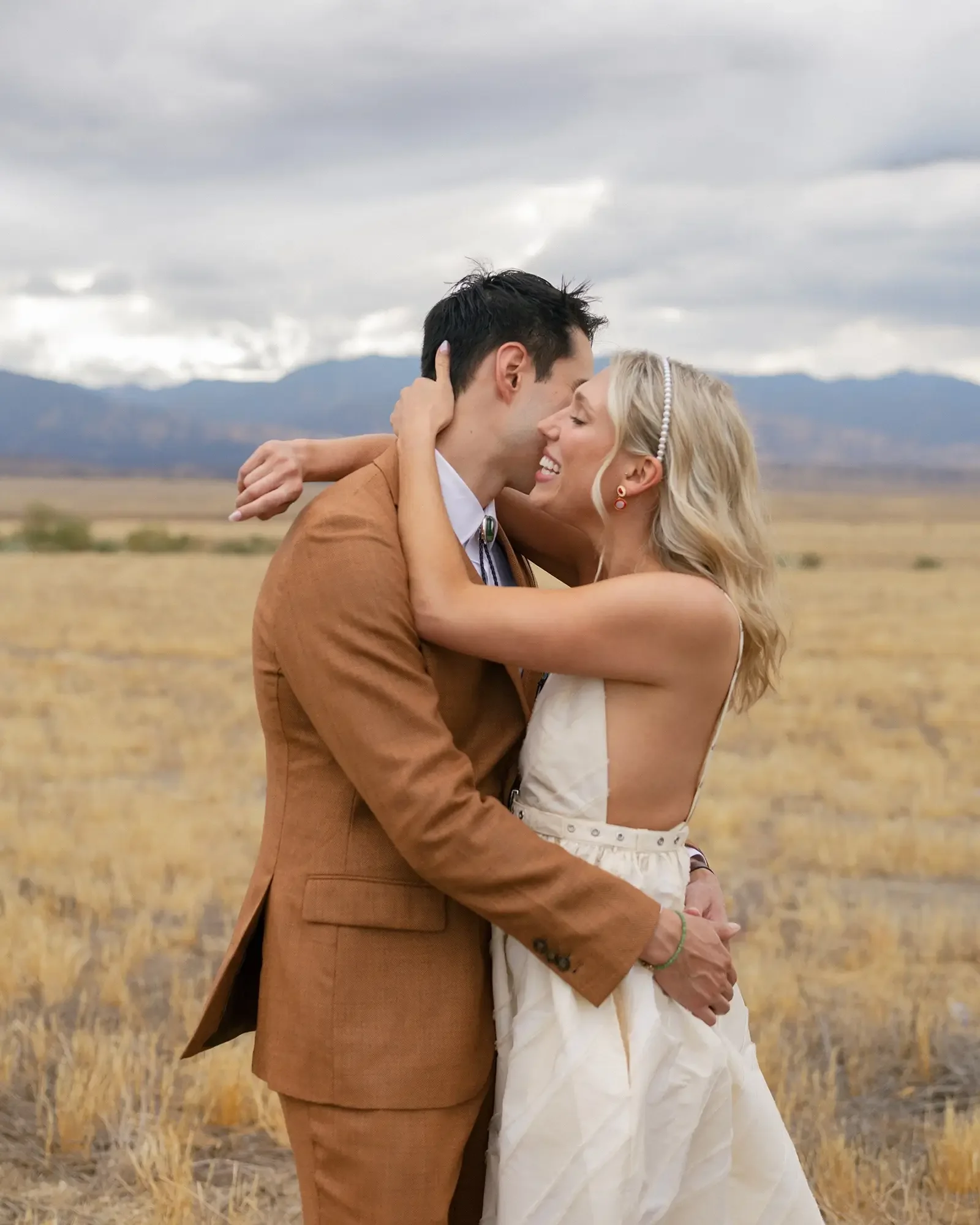 A couple embracing outdoors in a field with mountains and cloudy sky in the background.