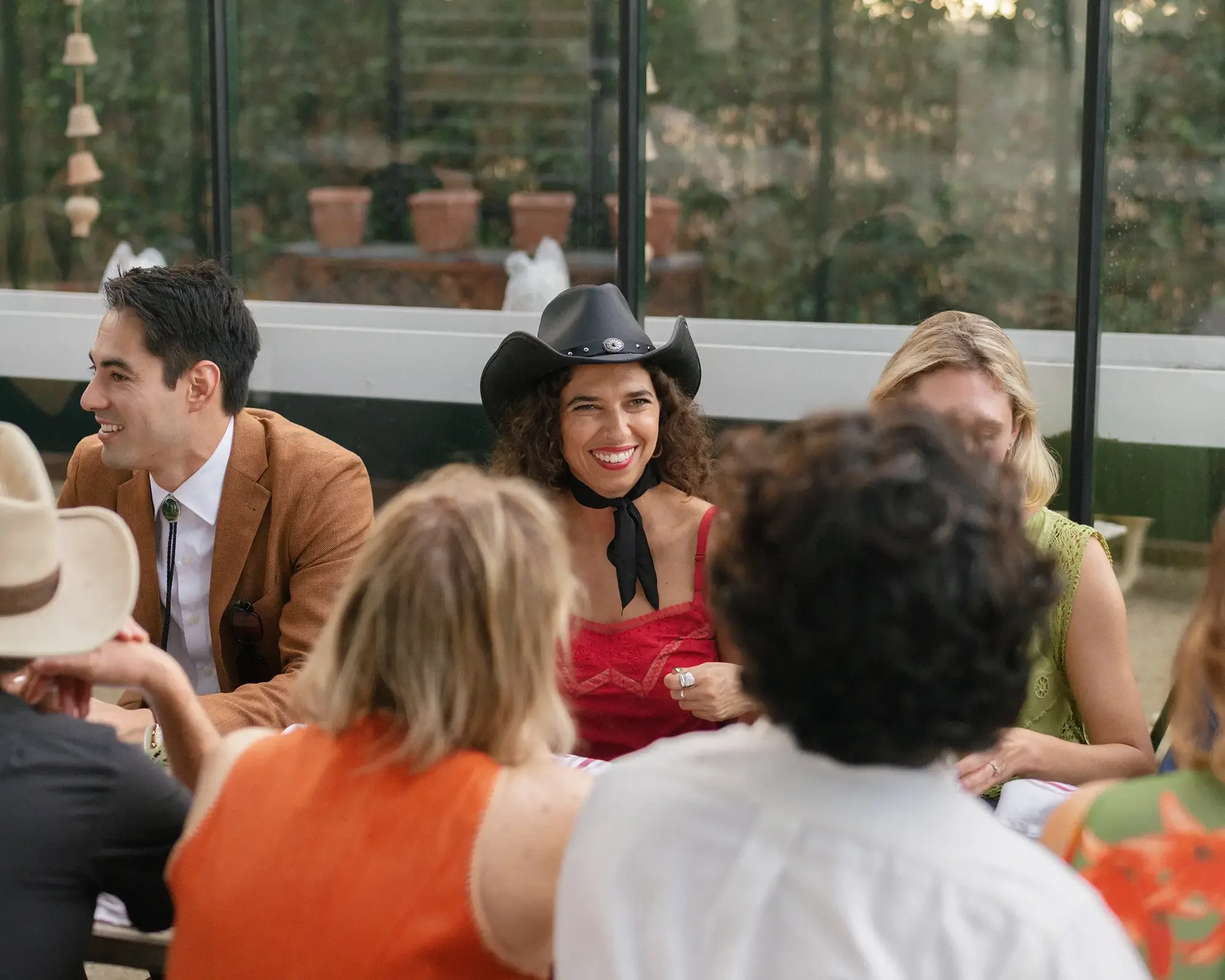 Rehearsal dinner guests sitting at a table, smiling and talking, with a woman wearing a black cowboy hat and red dress as a focal point.