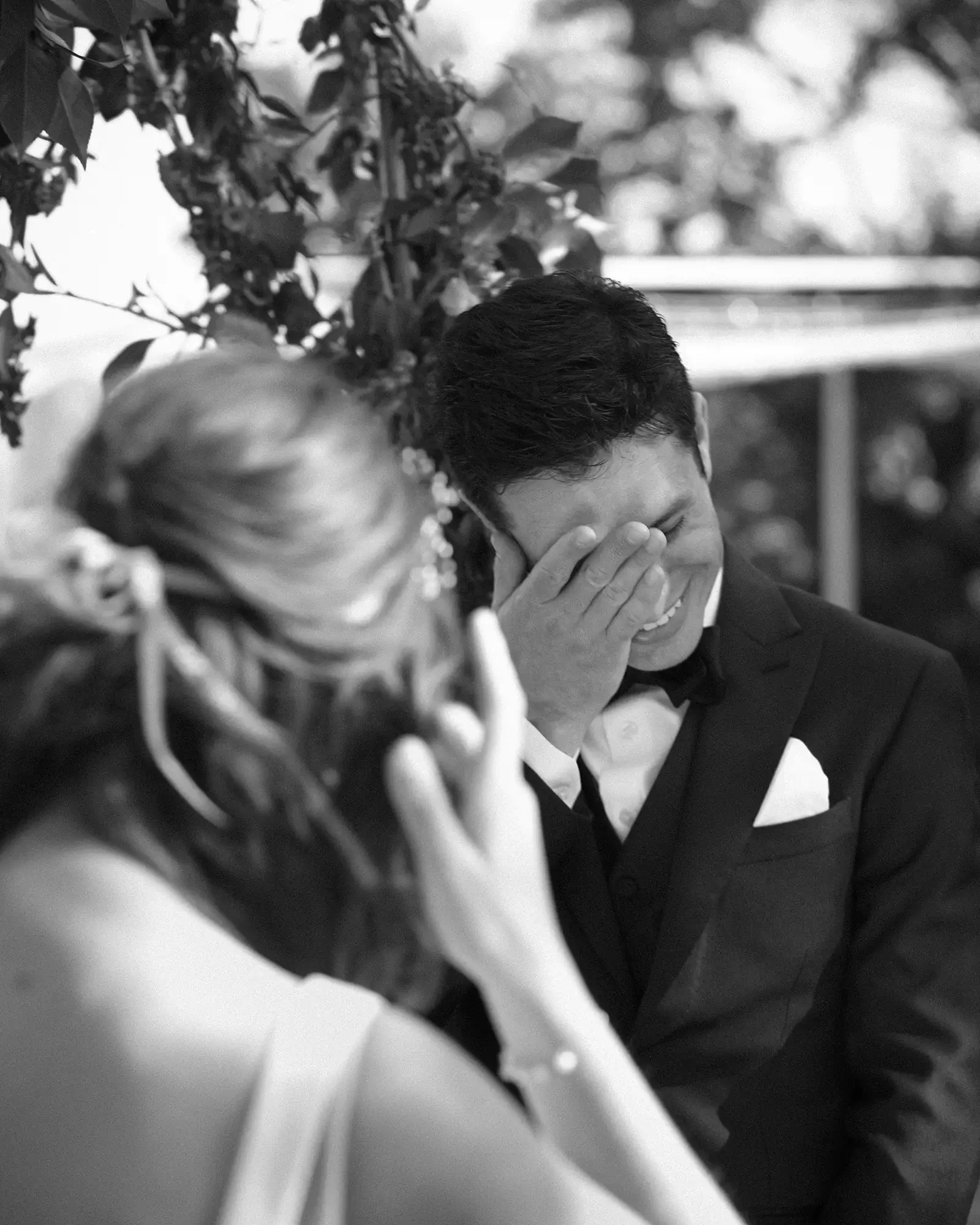 A groom in a tuxedo covering his face with his hand, emotional, and a bride with glasses and earrings in the foreground touching her face, indicating a tender moment.