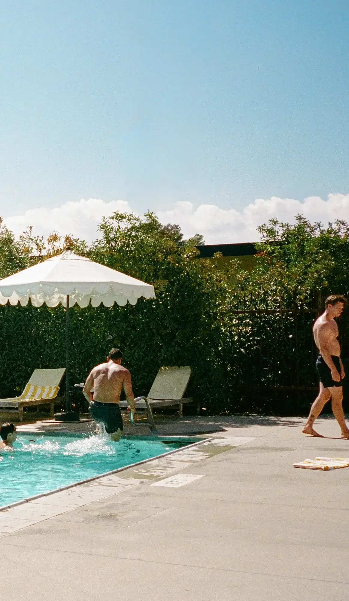 People swimming and playing by a pool outdoors on a sunny day, with a white umbrella and lounge chairs visible.