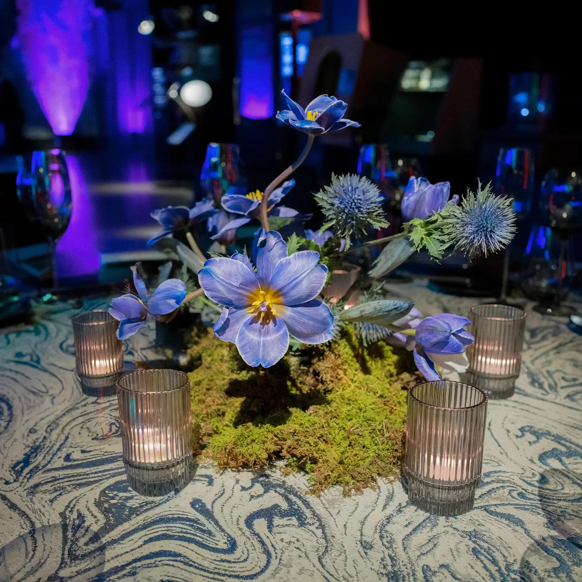 Elegant table centerpiece with blue flowers and moss, surrounded by small candles in glass holders, set on a patterned tablecloth in a dimly lit environment with purple and blue lighting.