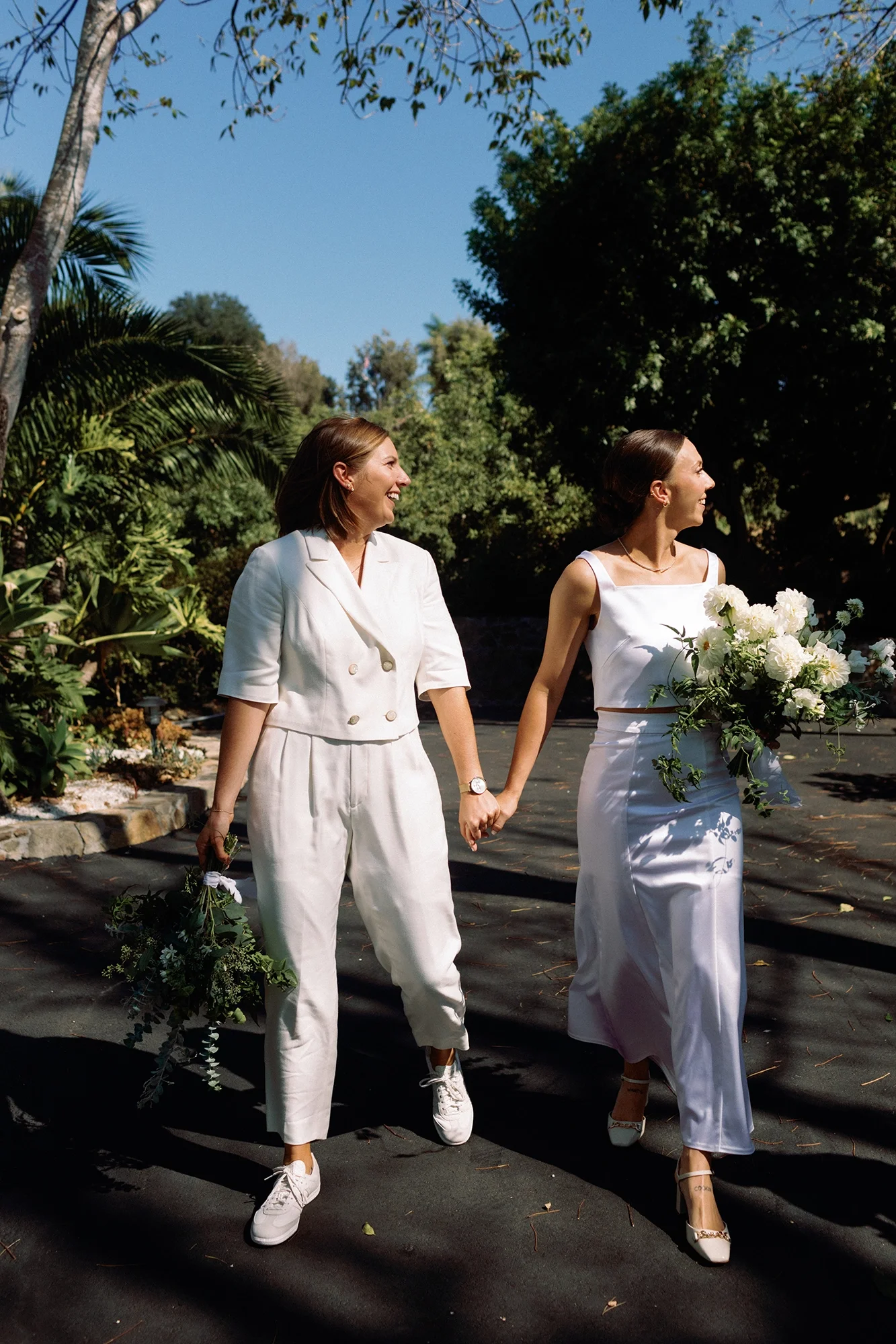 Two brides in their wedding day outfits holding hands and bouquets, smiling, walking outdoors on a sunny day in a lush, green park.