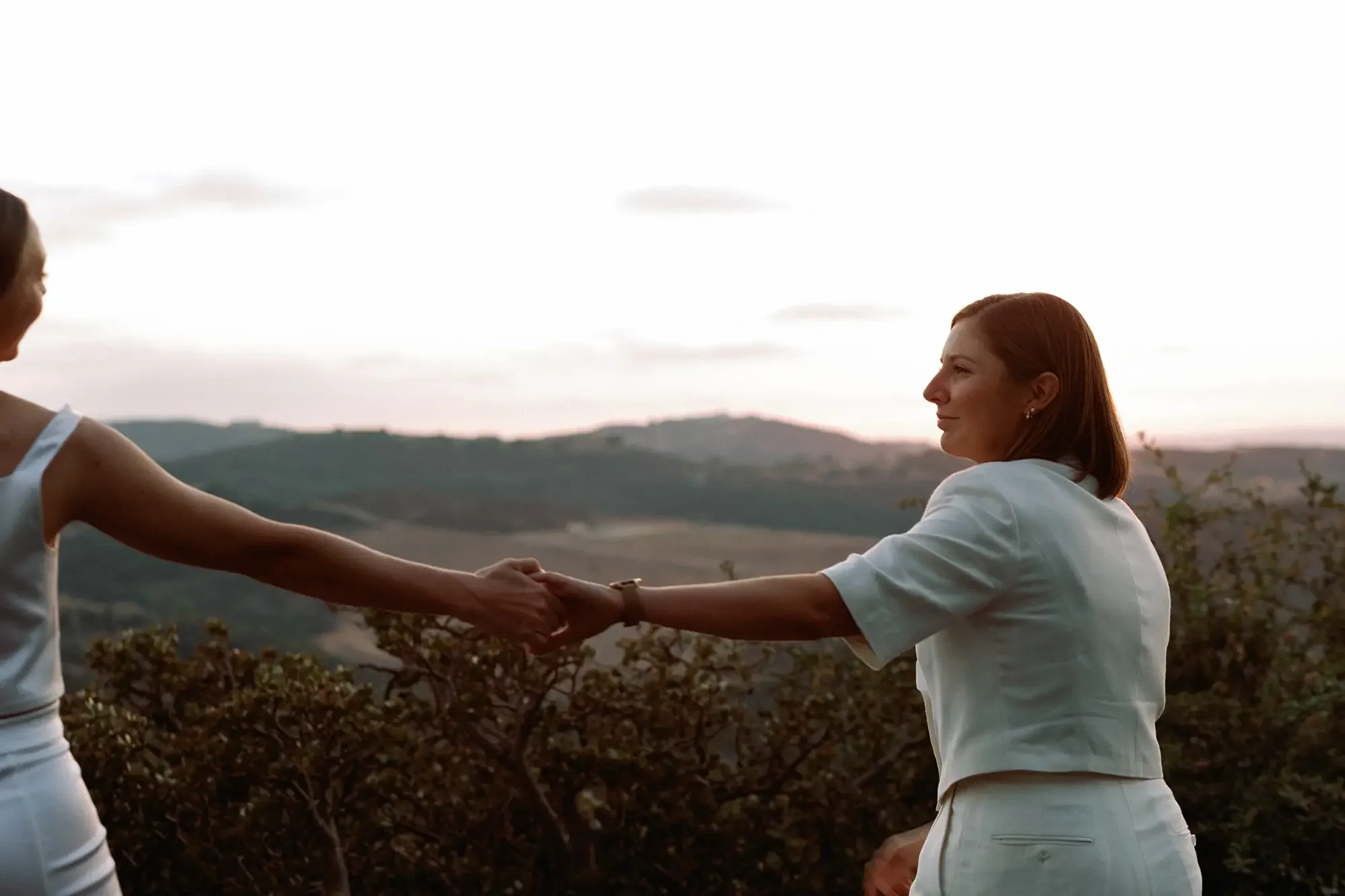Two brides holding hands outdoors during sunset, with rolling hills in the background.
