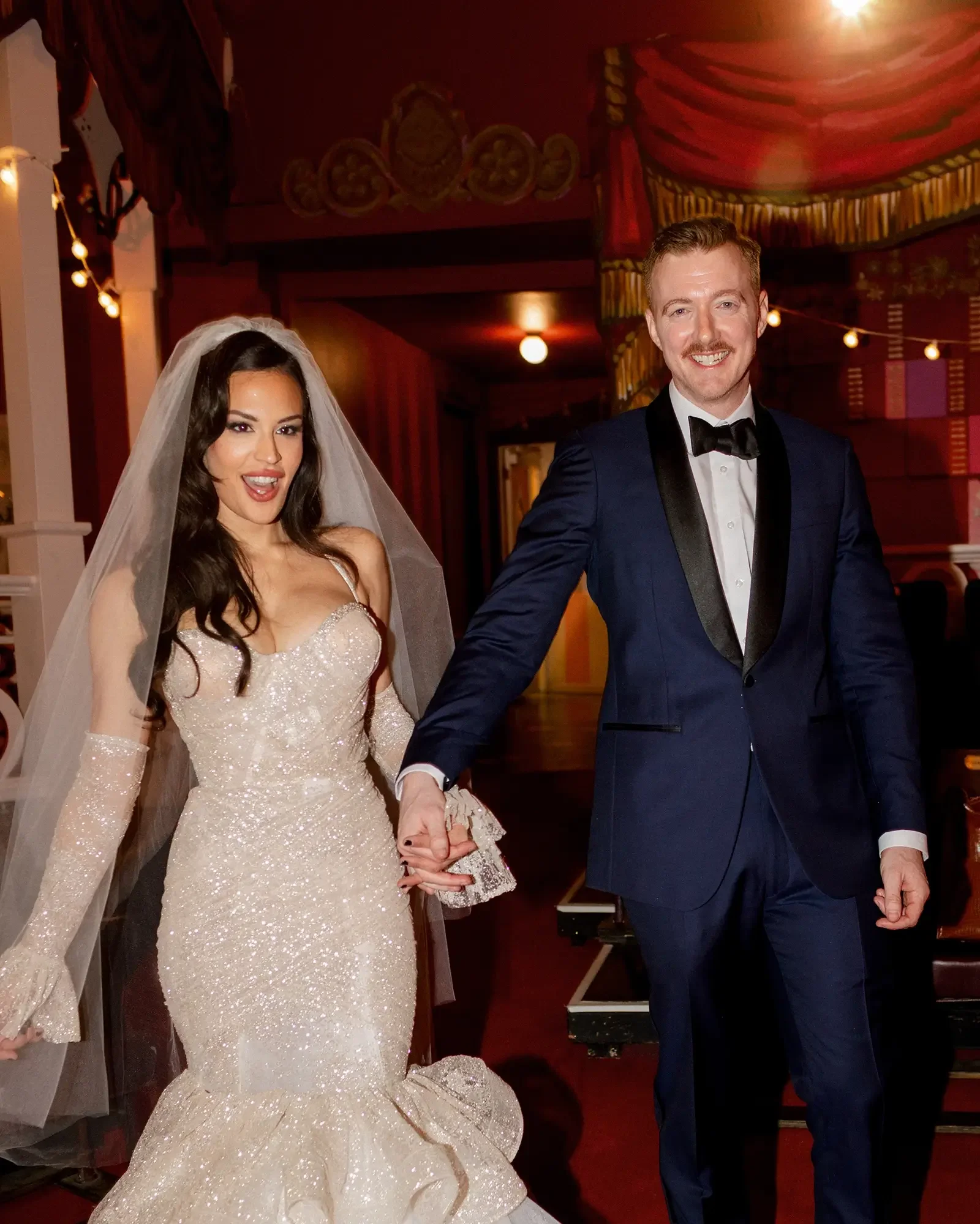 A bride and groom holding hands and smiling indoors at a wedding reception, with the bride in a white, sparkly wedding dress and the groom in a dark tuxedo with a black bow tie.