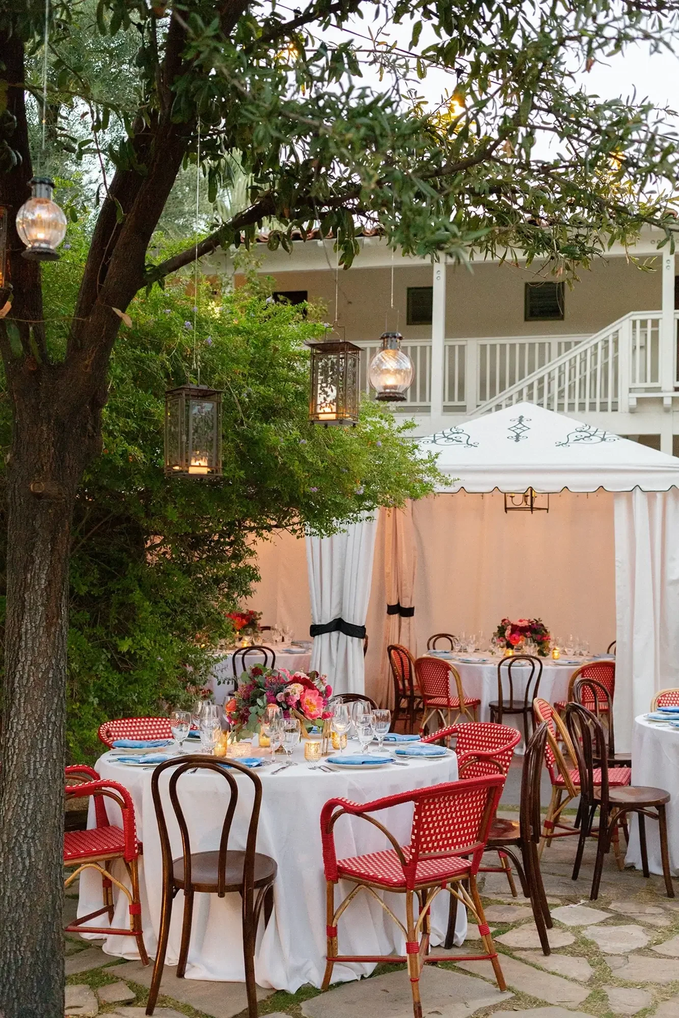 Outdoor wedding with round tables, white tablecloths, floral centerpieces, and various chairs in front of a white tent, with trees and lanterns hanging overhead.