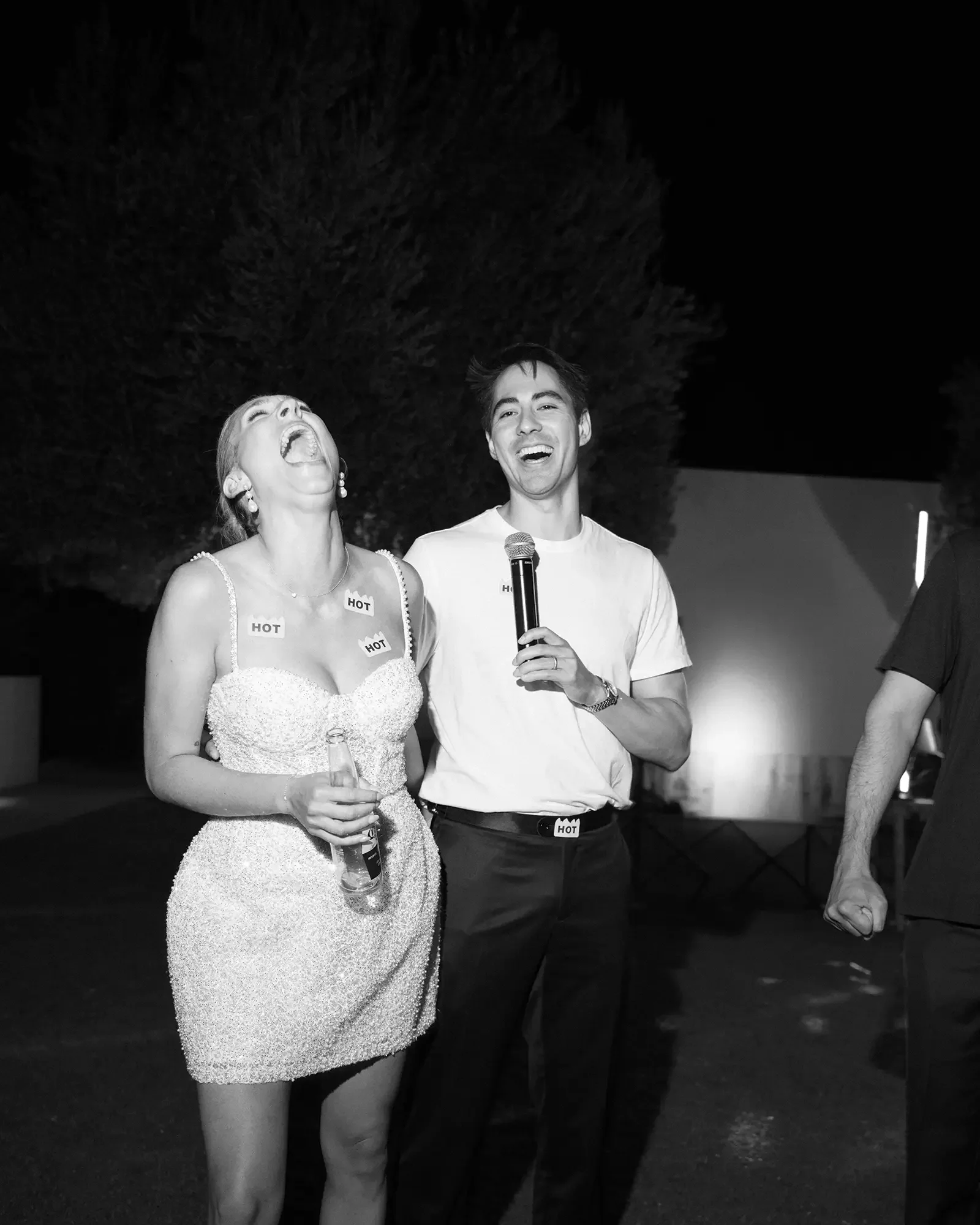 Black and white photo of a bride and groom laughing and enjoying themselves at the wedding reception at night. The woman is holding a bottle, and the man is holding a microphone. Both are wearing stickers that say 'HOT' on their shirts.