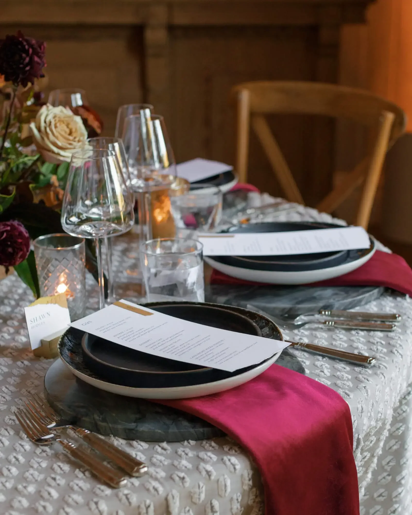 An elegant wedding dining table with black and white plates, pink cloth napkins, wine glasses, water glasses, candles, and a floral centerpiece with roses, on a white textured tablecloth in a cozy wooden interior.