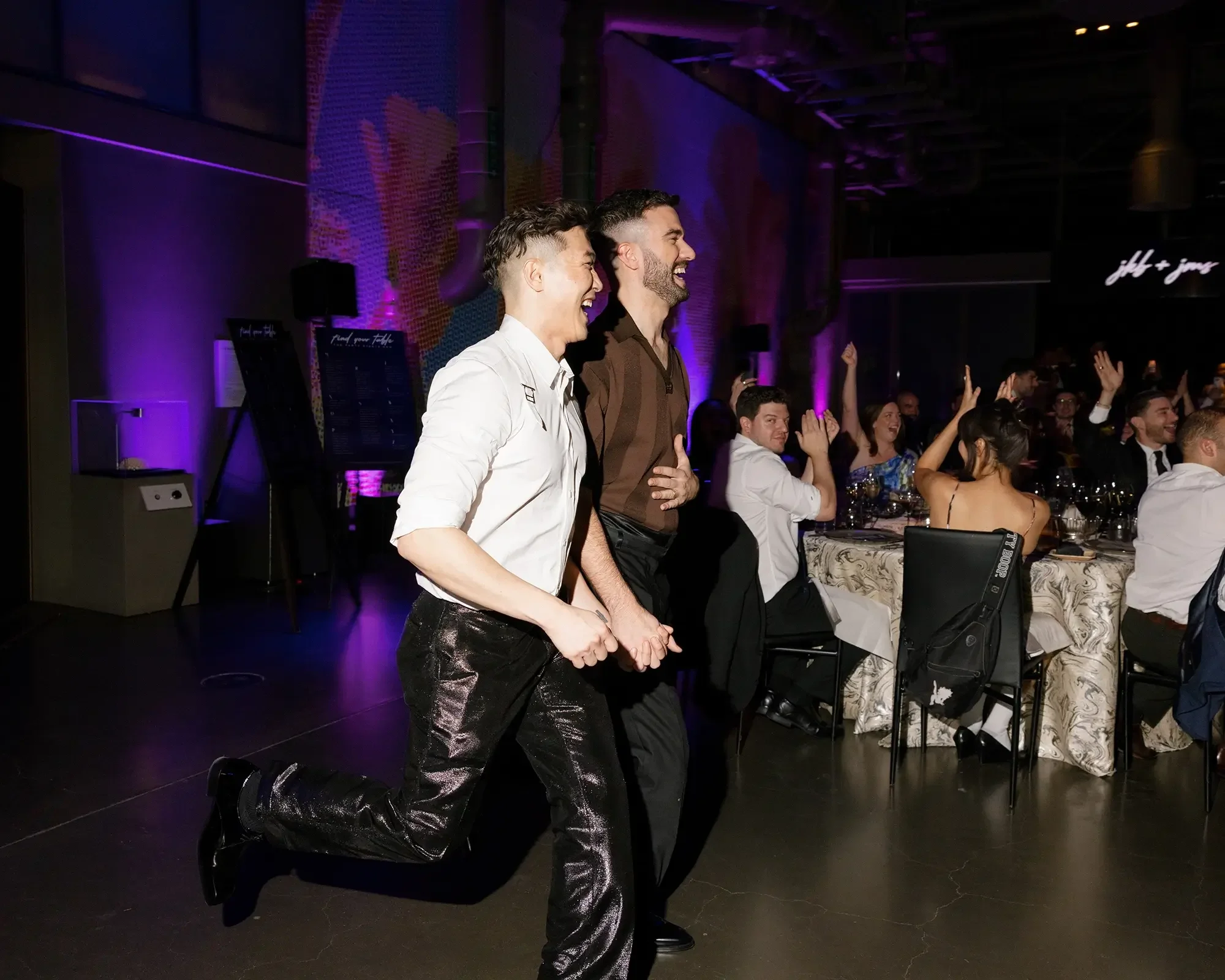 Two grooms' grand entrance to their wedding reception, holding hands while laughing at a lively party or celebration, with other people seated at tables in the background.
