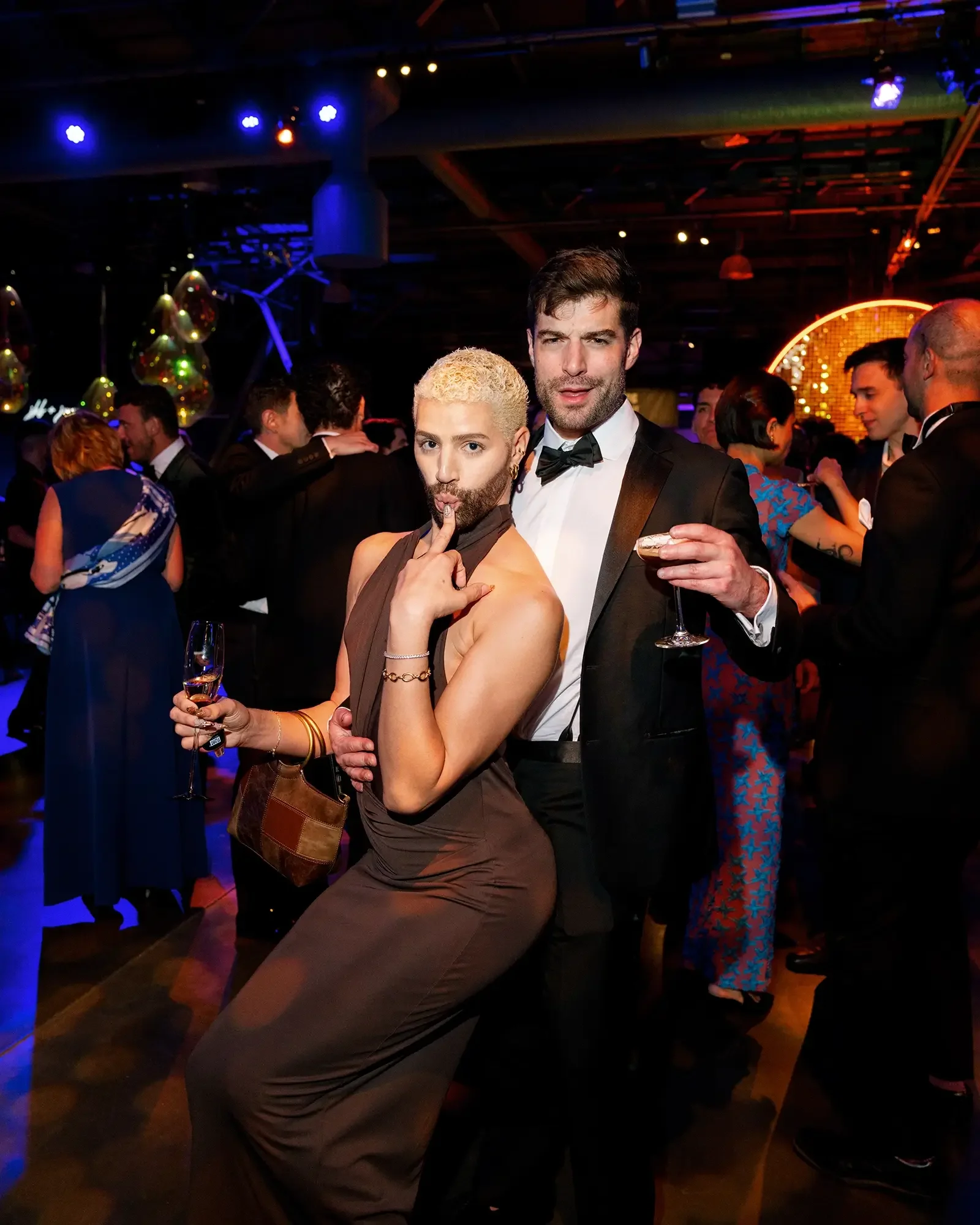Two people dressed in formal attire on the dance floor at a wedding, with one is holding a glass of champagne and the other with finger to their lips, standing in a lively, decorated setting with others in the background.