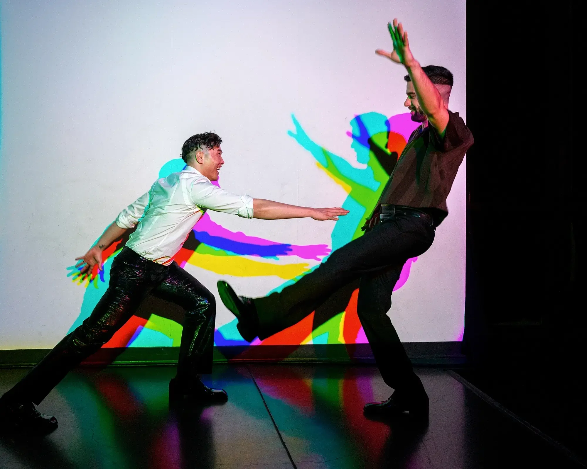 Two grooms dancing with colorful shadows projected onto a plain white background, one reaching out and the other kicking.