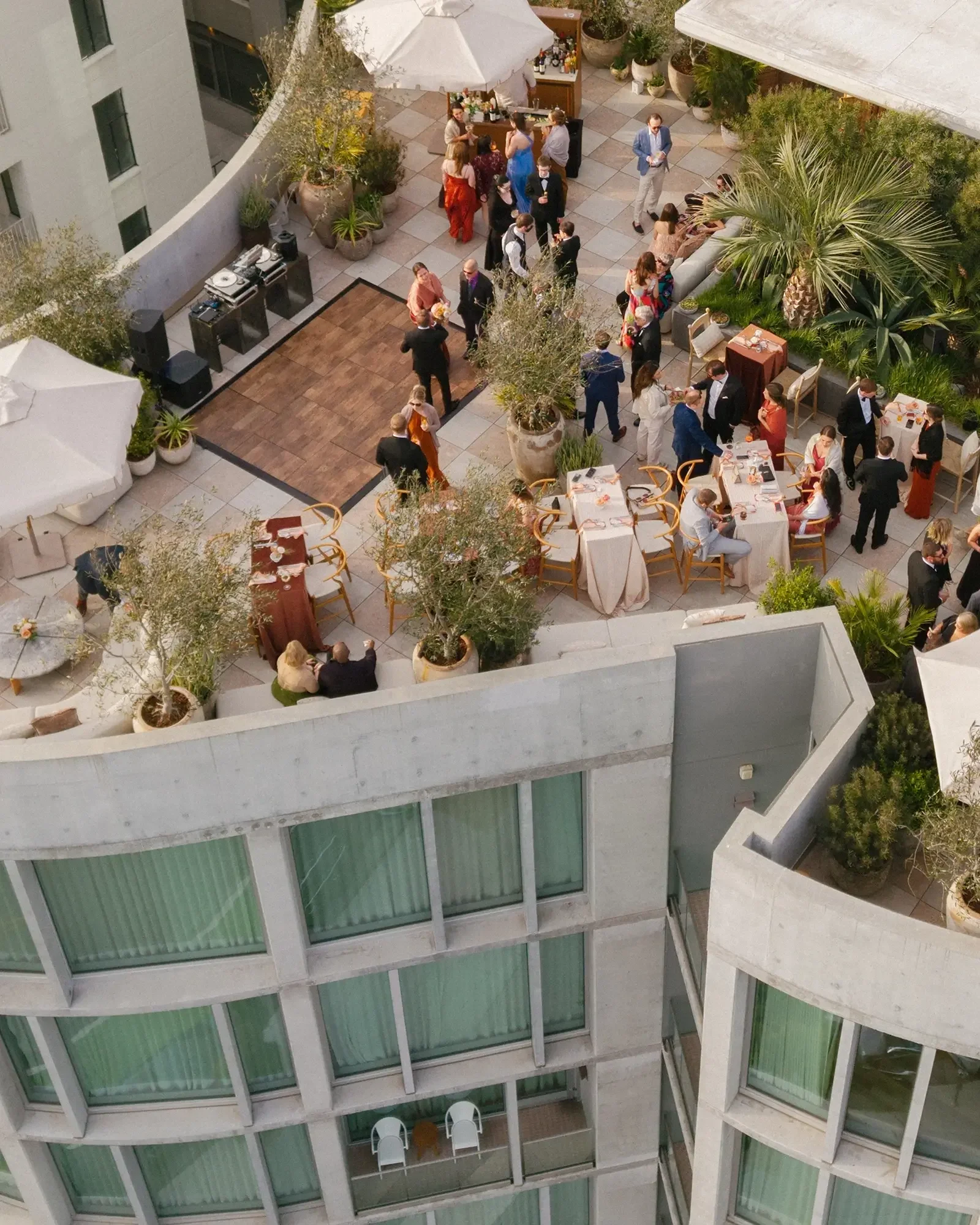A rooftop cocktail hour with people socializing, eating, and drinking. There are tables with white tablecloths, potted plants, and a dance floor area, as well as an outdoor bar .