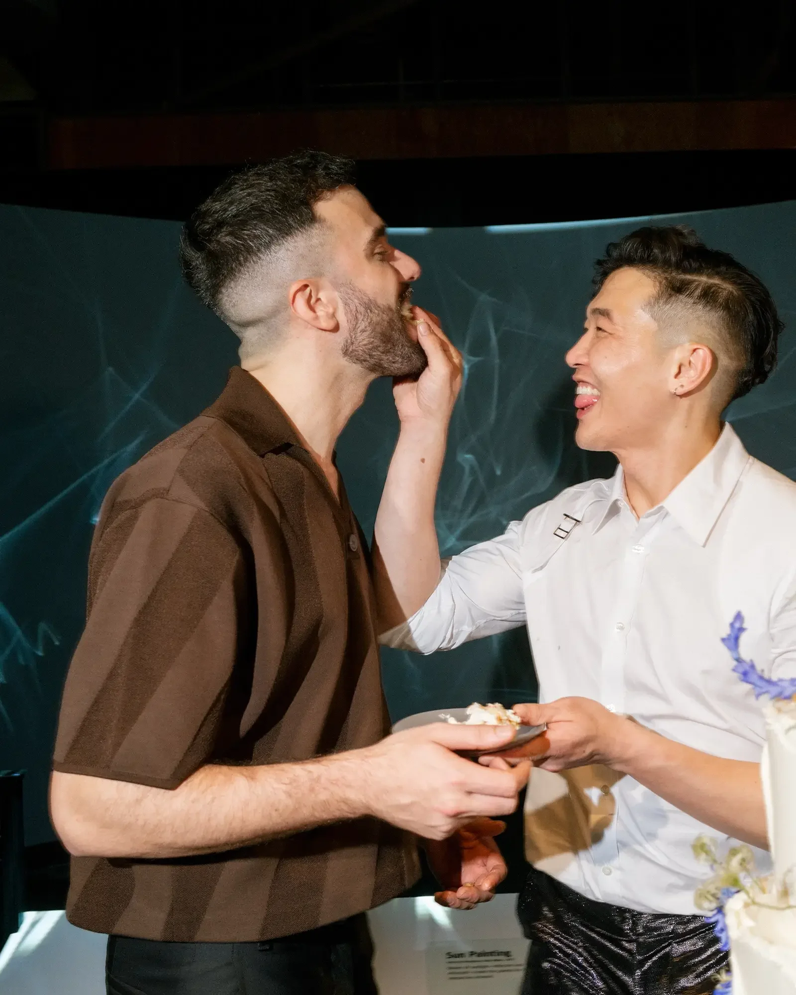 Two grooms smiling and sharing a piece of cake, one feeding the other during their wedding.