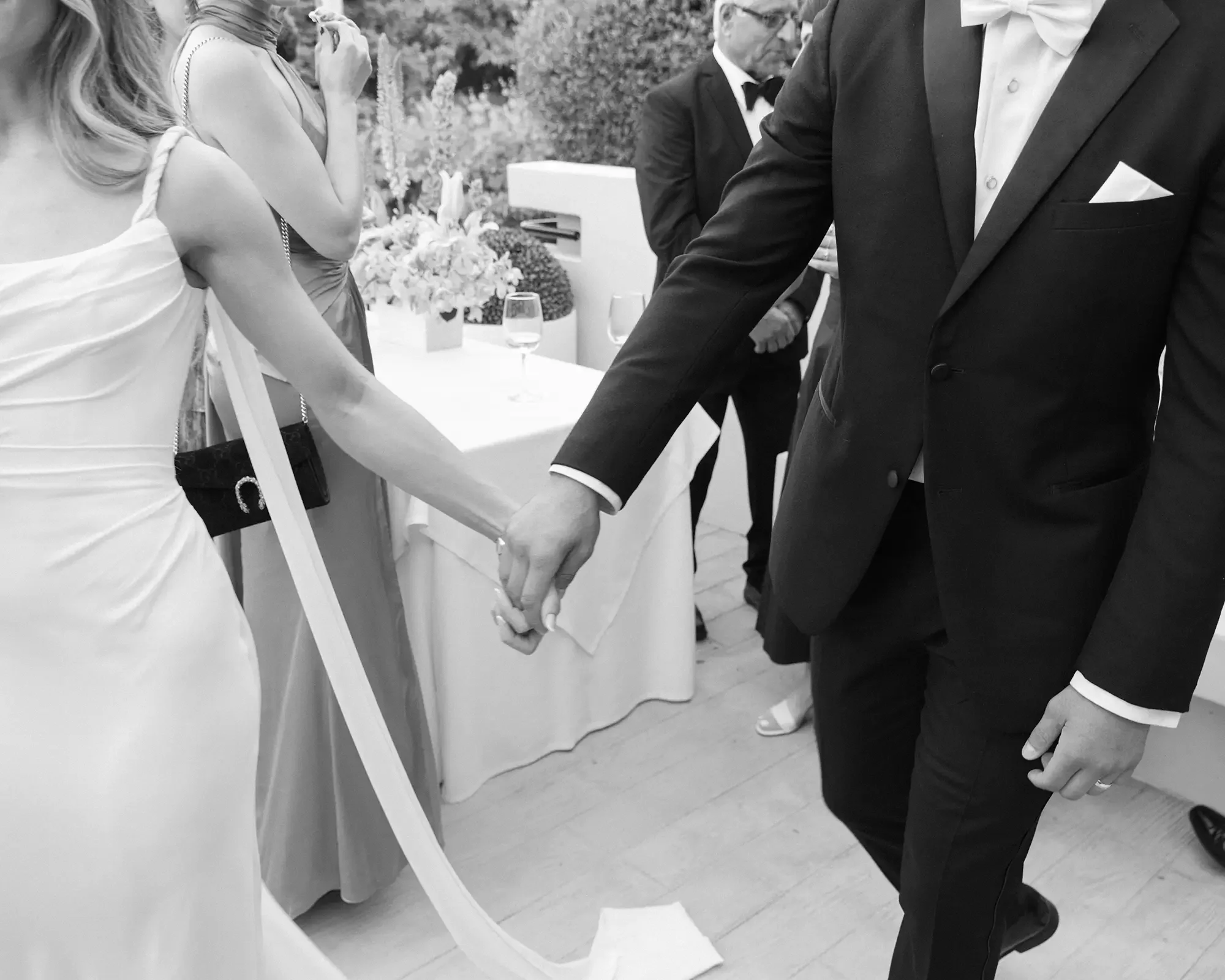 A bride and groom holding hands at their wedding with other guests in the background, dressed in evening wear, near a table with flowers and drinks.