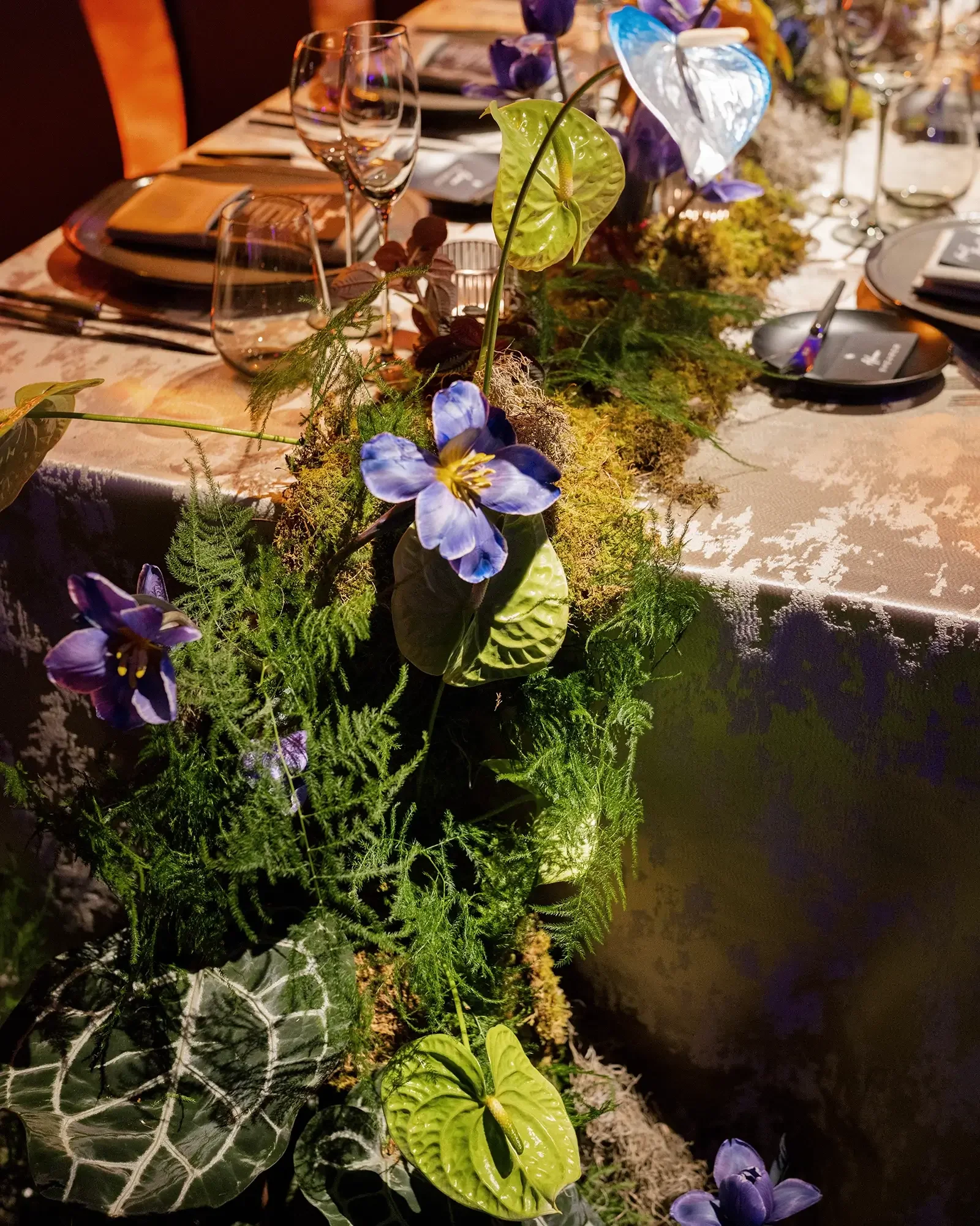 Wedding reception table decorated with a floral centerpiece featuring blue flowers and green leaves, with plates, glasses, and utensils arranged for a meal.