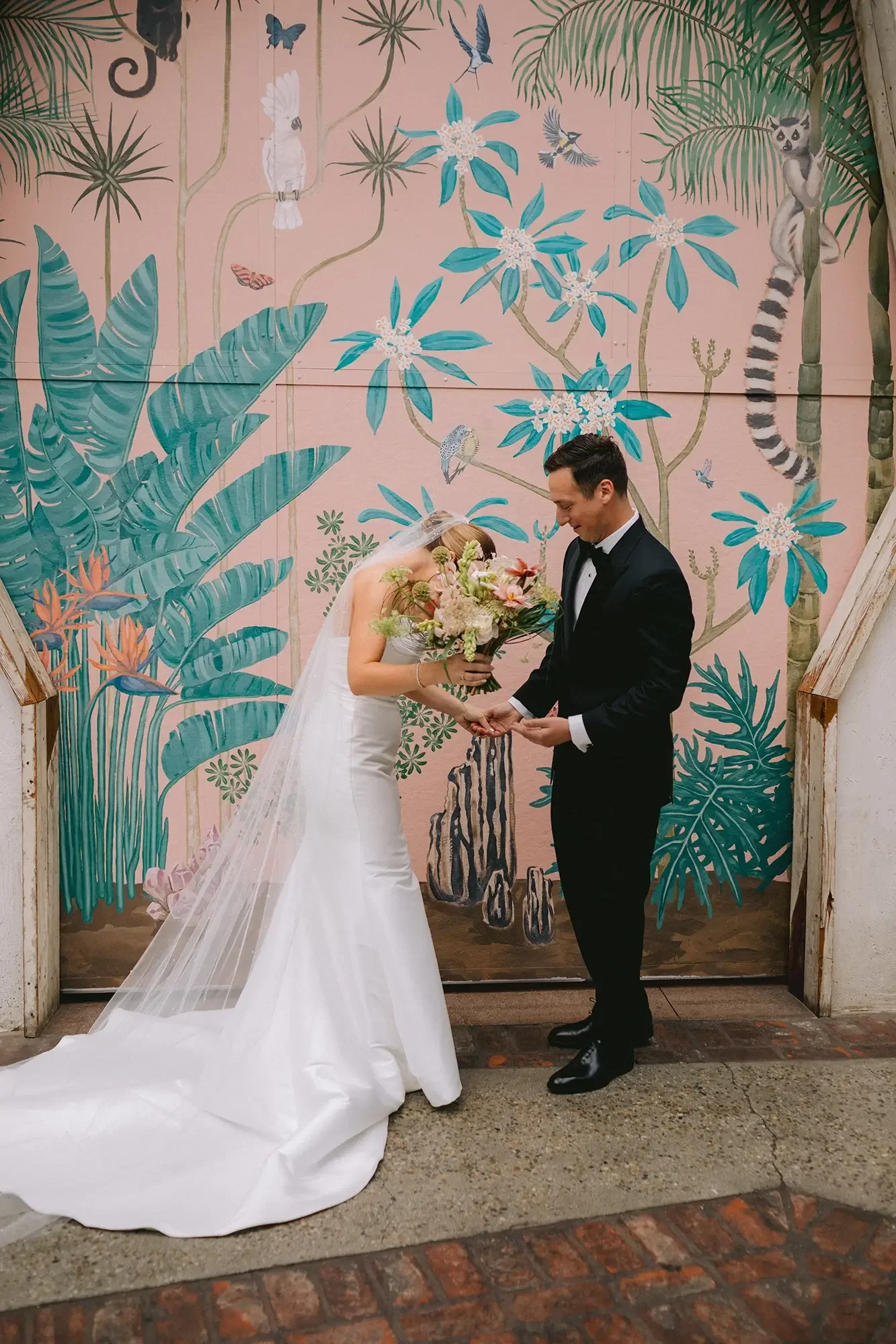 A bride and groom holding hands and bowing their heads in front of a colorful jungle-themed mural with birds, monkeys, and tropical plants, during a wedding ceremony.