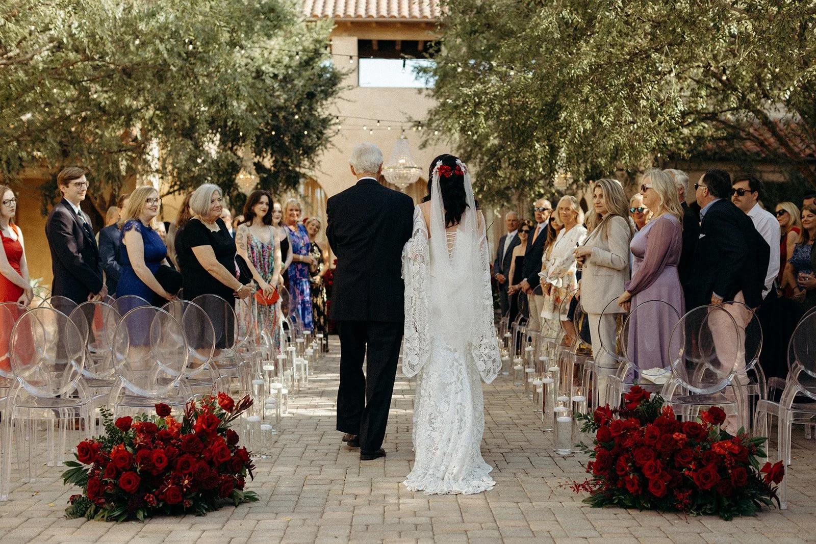 A bride in a white lace wedding dress and veil walking down the aisle on her wedding day, flanked by guests on both sides, outdoors under large leafy trees with a chandelier hanging above.