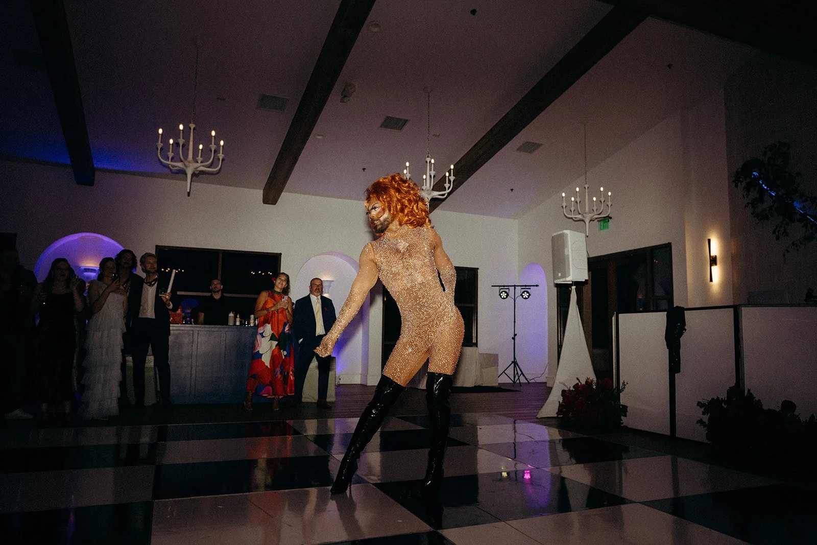 A performer in a sheer, sparkling outfit with black thigh-high boots dancing on a checkered dance floor at an indoor event, with guests watching in the background.