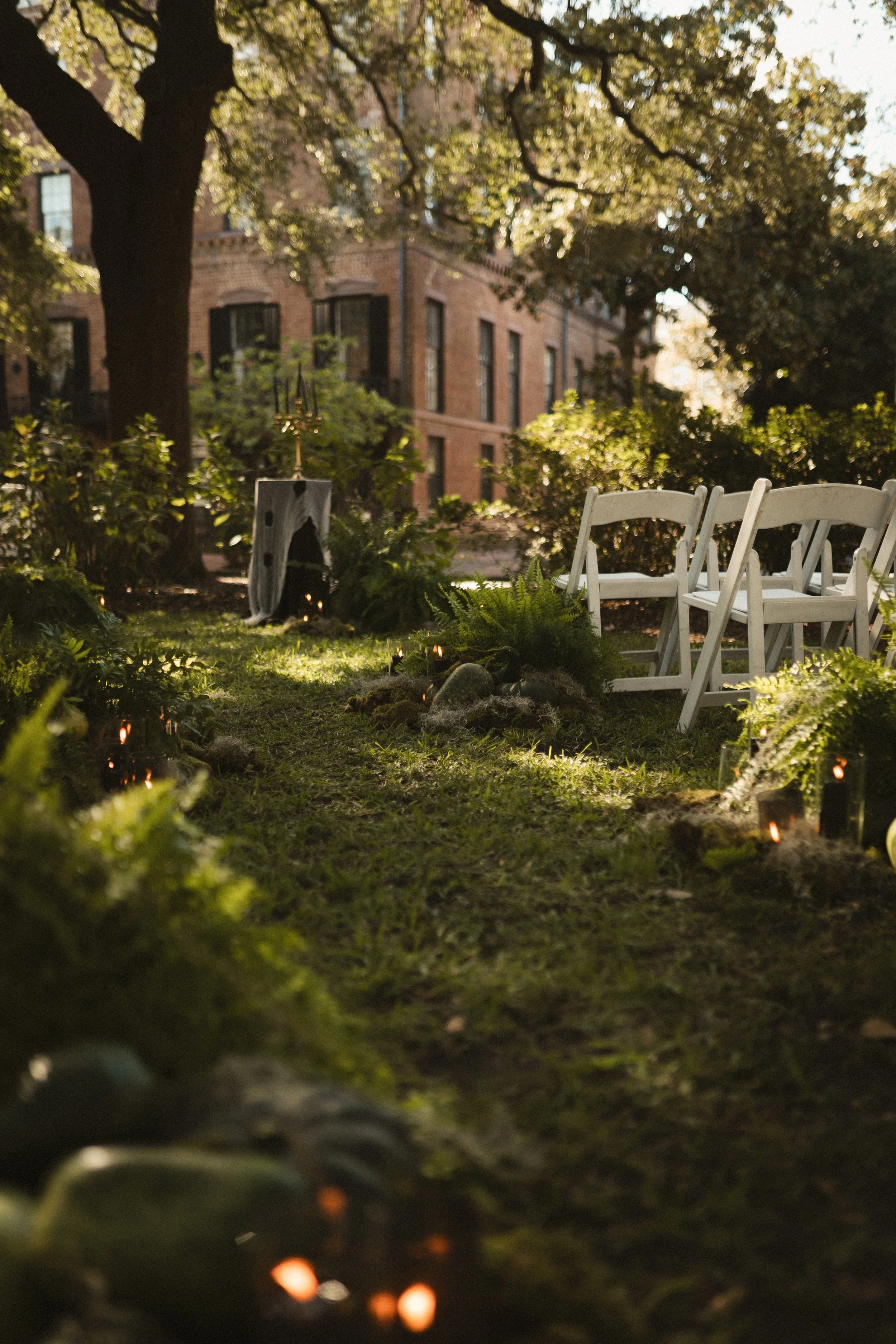 Outdoor garden venue with white folding chairs, lush trees, greenery, and a brick building in the background