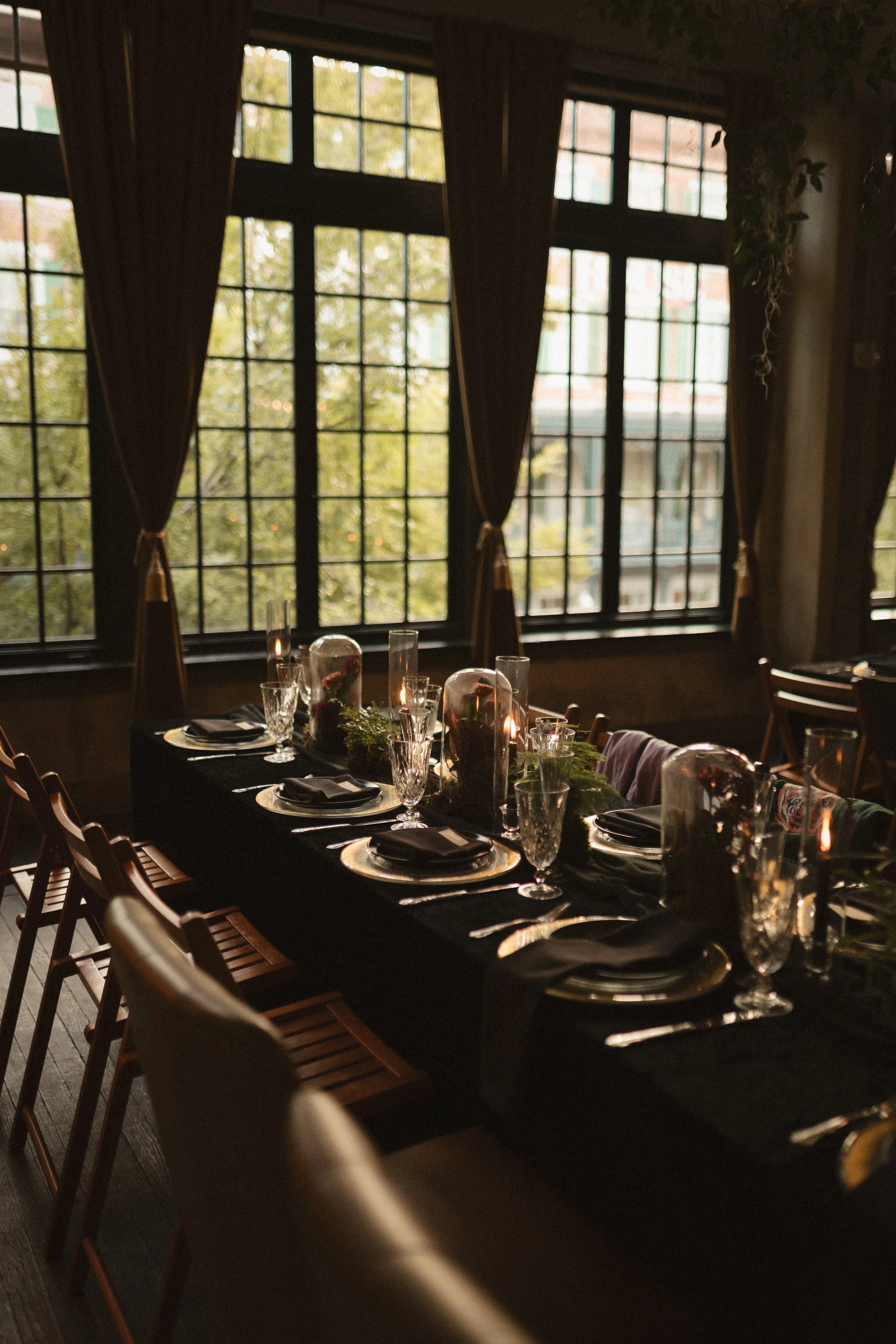 Elegant restaurant dining table set with black plates, glassware, candles, and greenery beside large windows with dark curtains