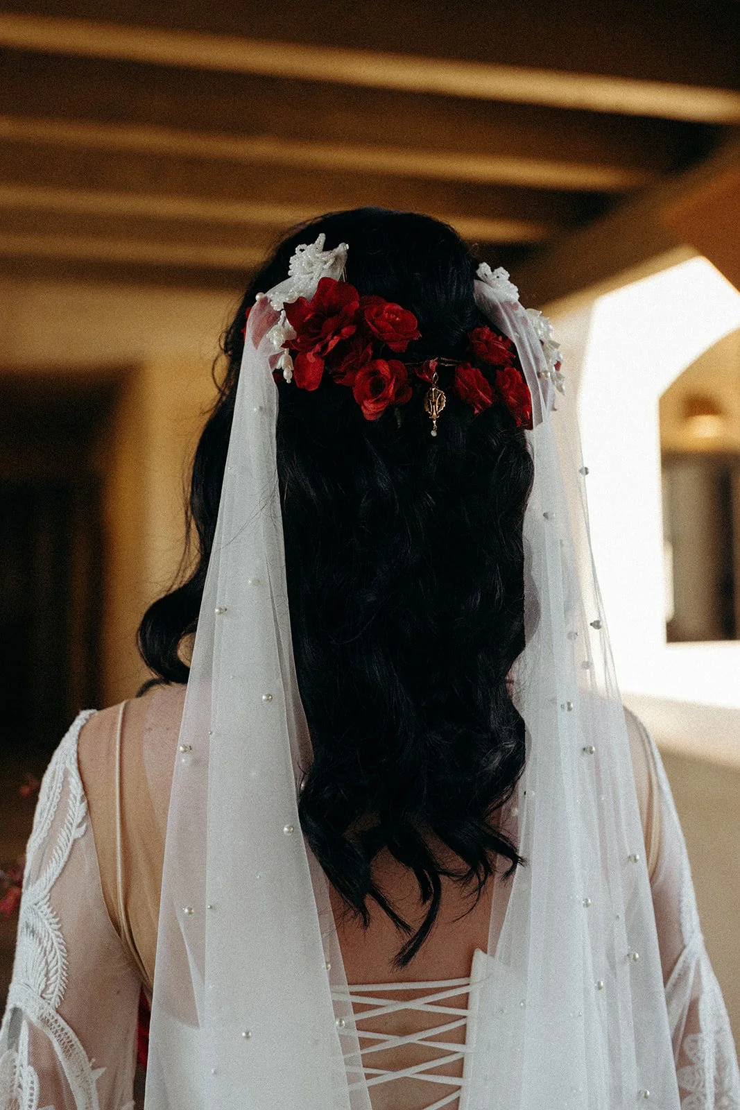 Back view of a woman with long black wavy hair, wearing a white veil decorated with small pearls and red flowers, and a beige dress with lace details.