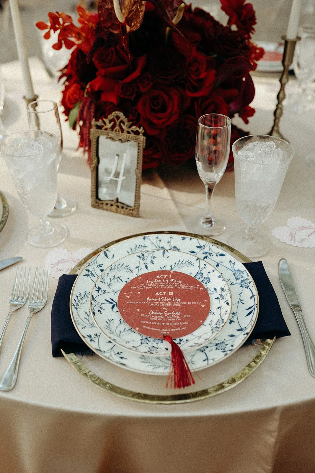 A dining table set with elegant plates, silverware, and glasses with ice. There is a large red floral centerpiece with roses and other flowers. A small framed table number '4' is positioned among the flowers. The plate has a detailed blue floral patt