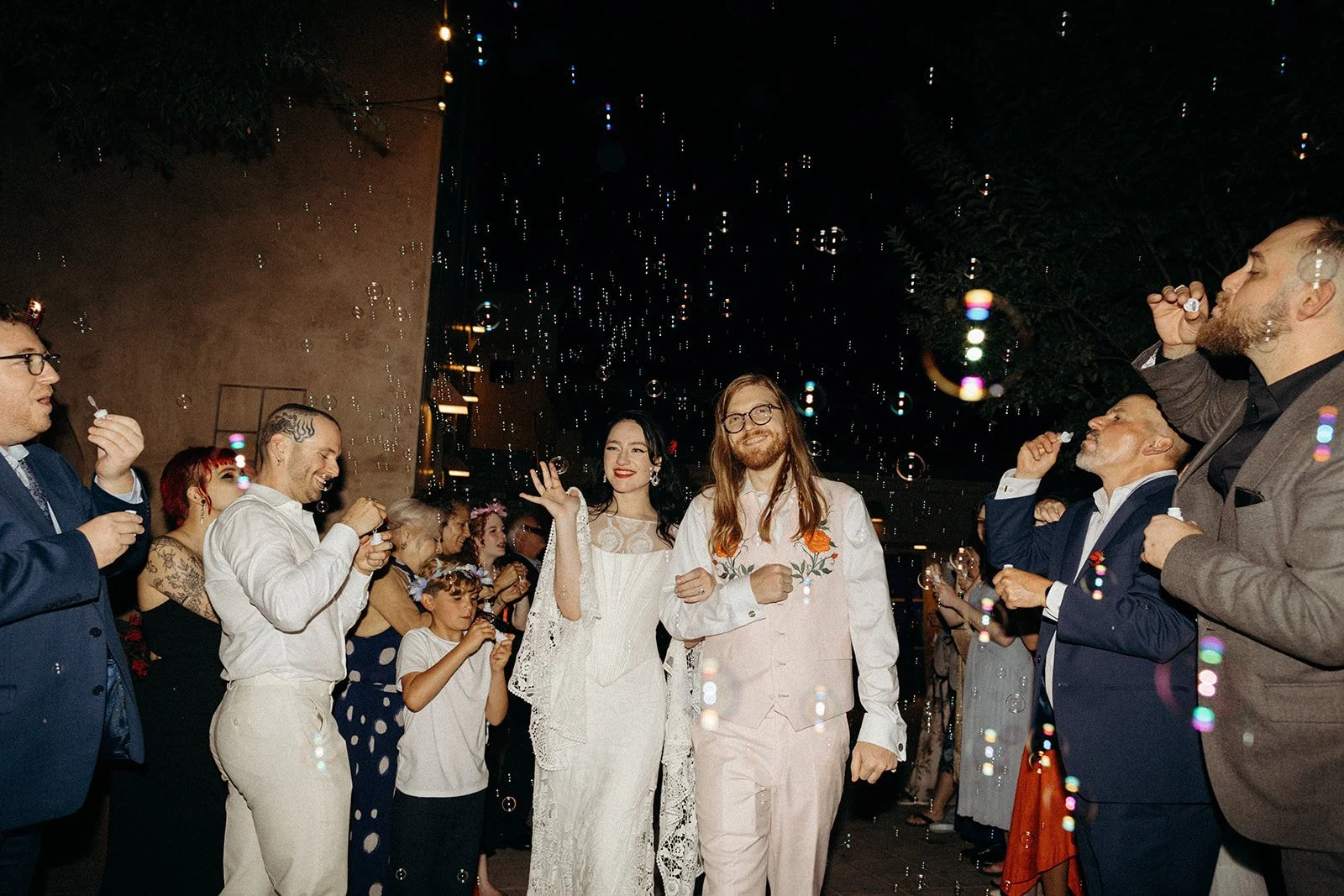 Couple walking down an aisle at a wedding reception, surrounded by guests blowing bubbles, celebration atmosphere, nighttime setting with lights.