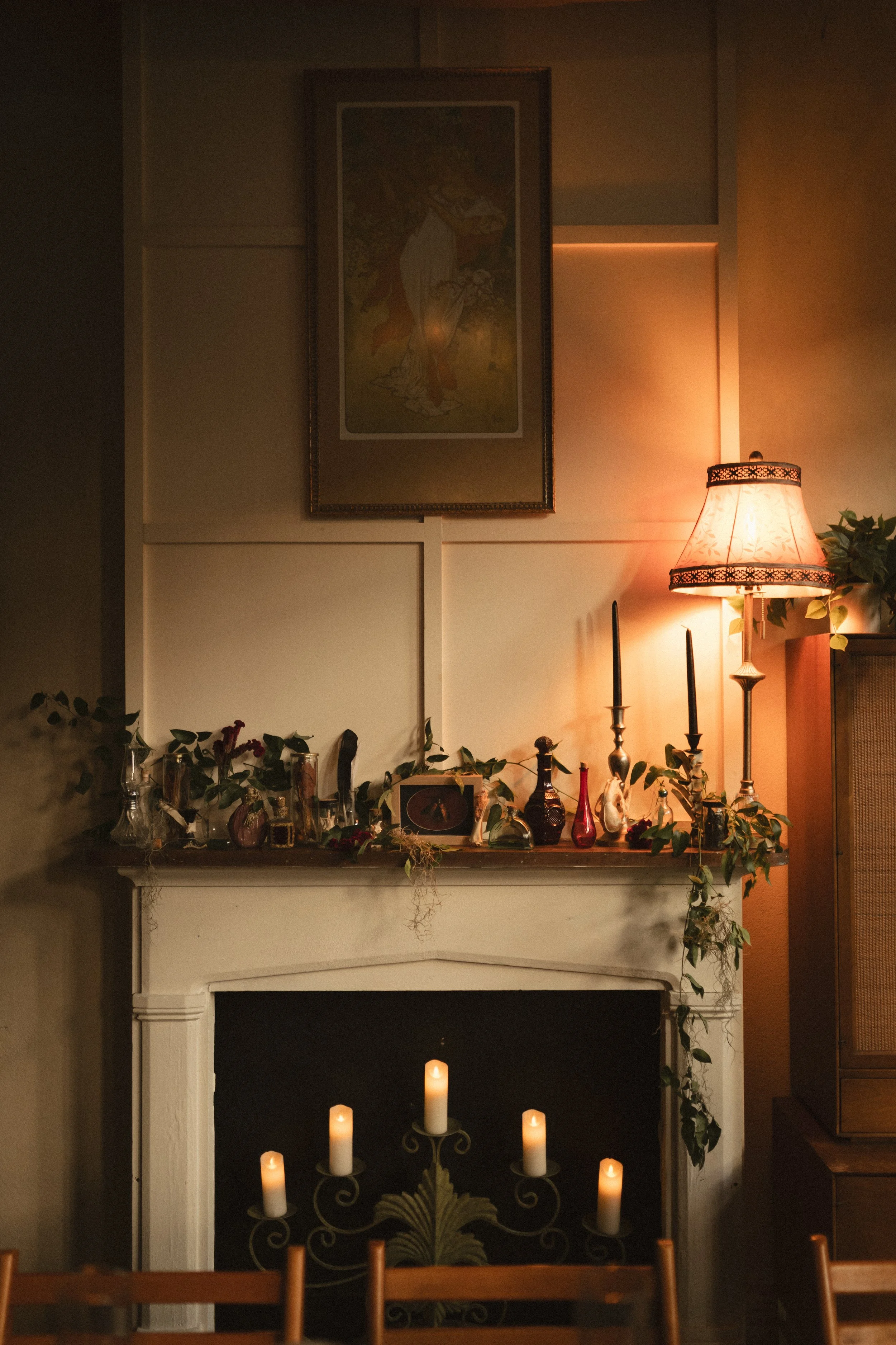 Cozy living room fireplace mantel decorated with plants, candles, framed artwork, and a glowing table lamp