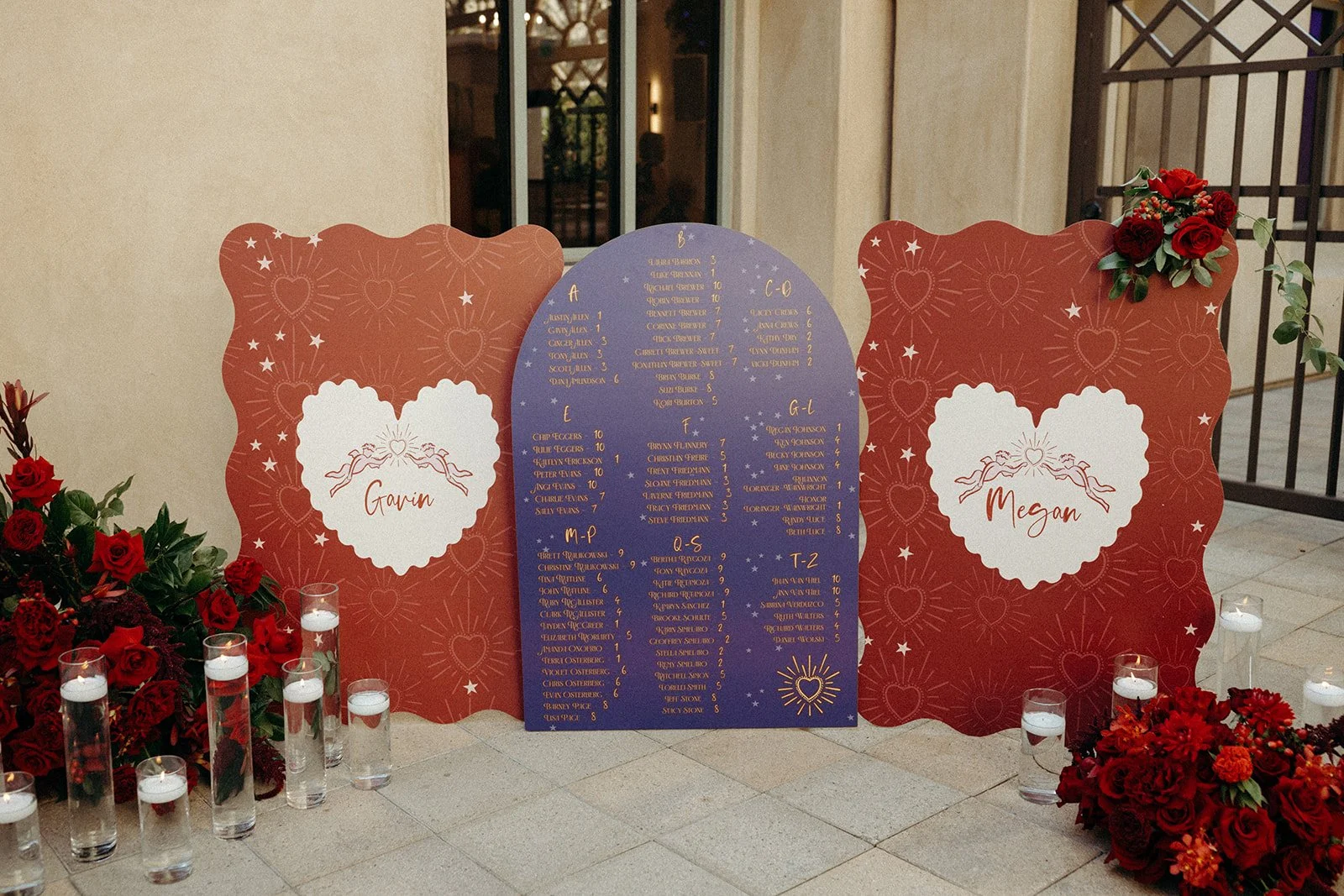 Wedding seating chart with two red decorative boards and a royal blue central board, surrounded by red roses and candles.