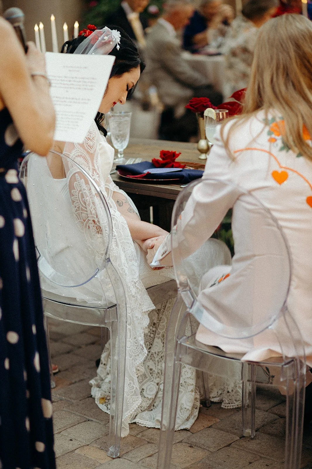 A bride with dark hair, wearing lace wedding dress and veil, holding hands with the groom wearing a white tux with floral designs, in a wedding ceremony setting with candles and red roses on the table.