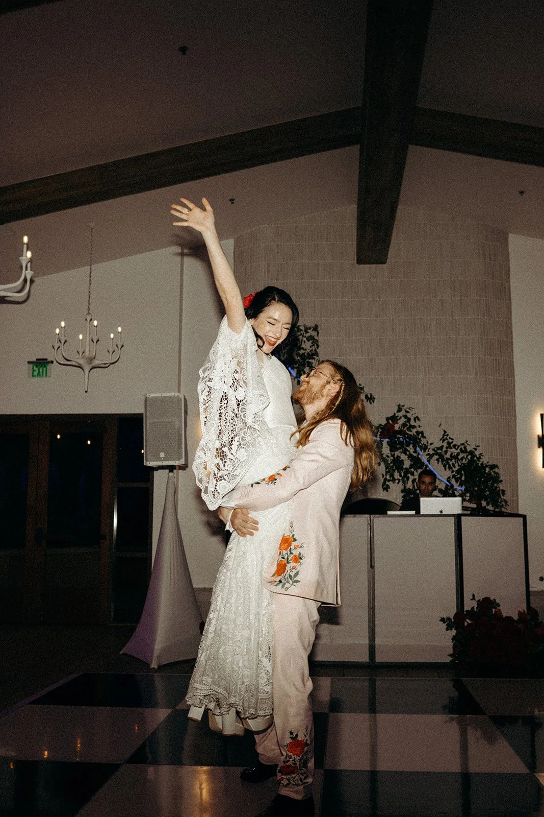 A woman in a white lace dress being lifted by a man in a cream-colored suit with embroidered floral patterns, both smiling and celebrating at a wedding reception.