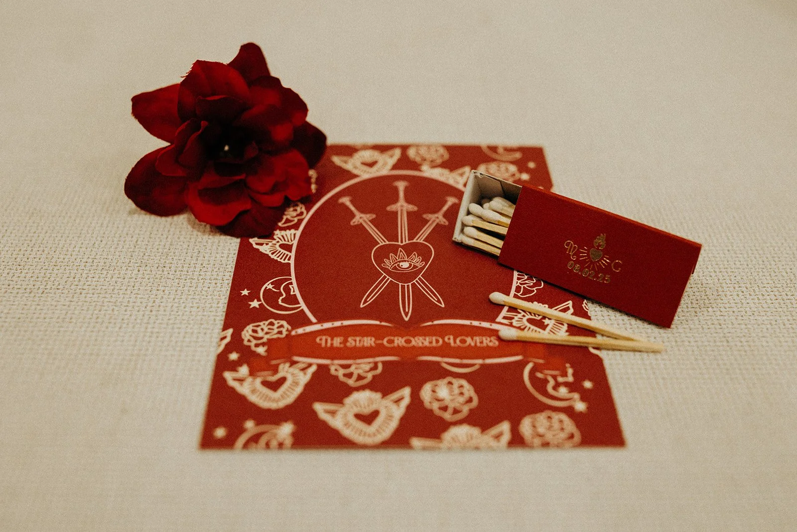 A red card with swords and heart graphic, a rose, a matchbox, and two matchsticks on a beige textured surface.