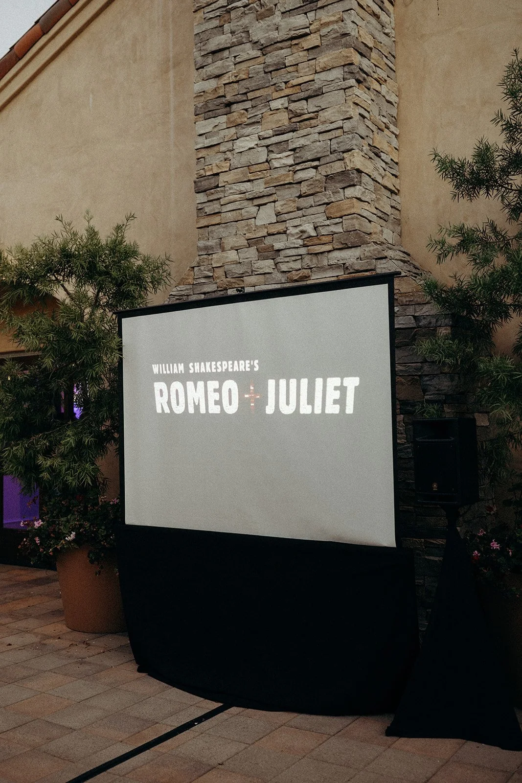 Projection screen displaying 'William Shakespeare's Romeo + Juliet' in an outdoor setting with stone and beige wall, potted plants, and trees around.