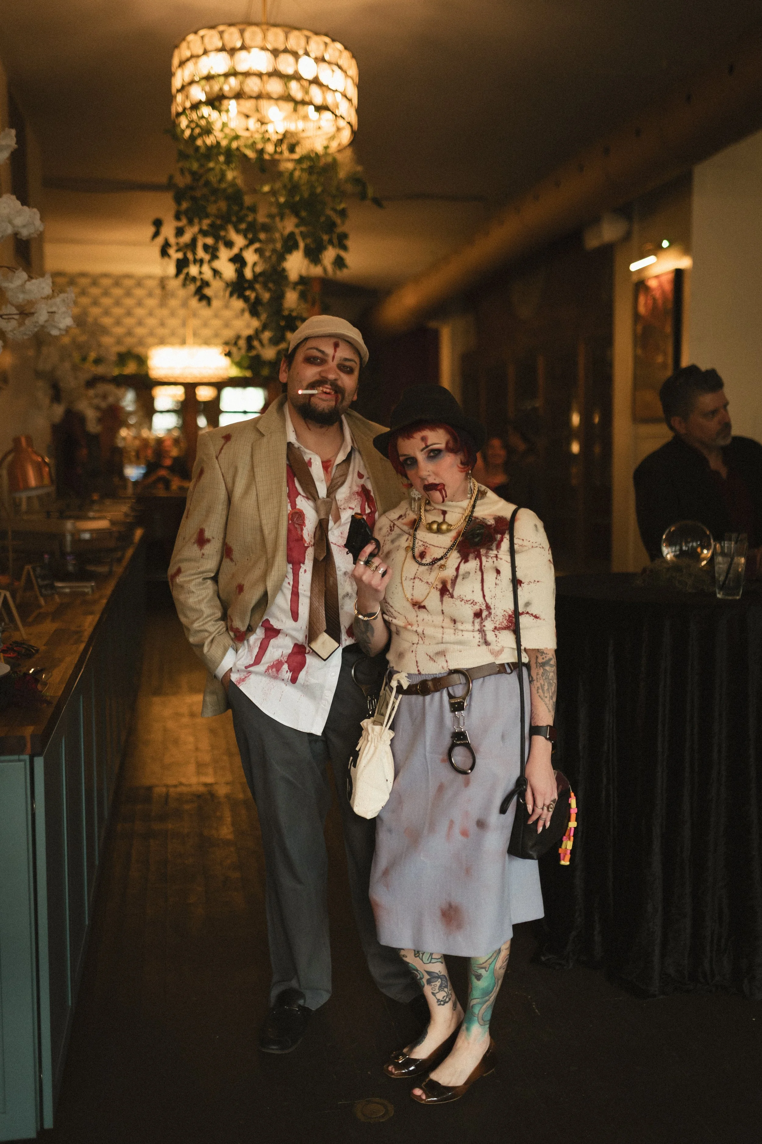 Two people in blood-stained zombie costumes pose at a dimly lit bar beneath a chandelier.