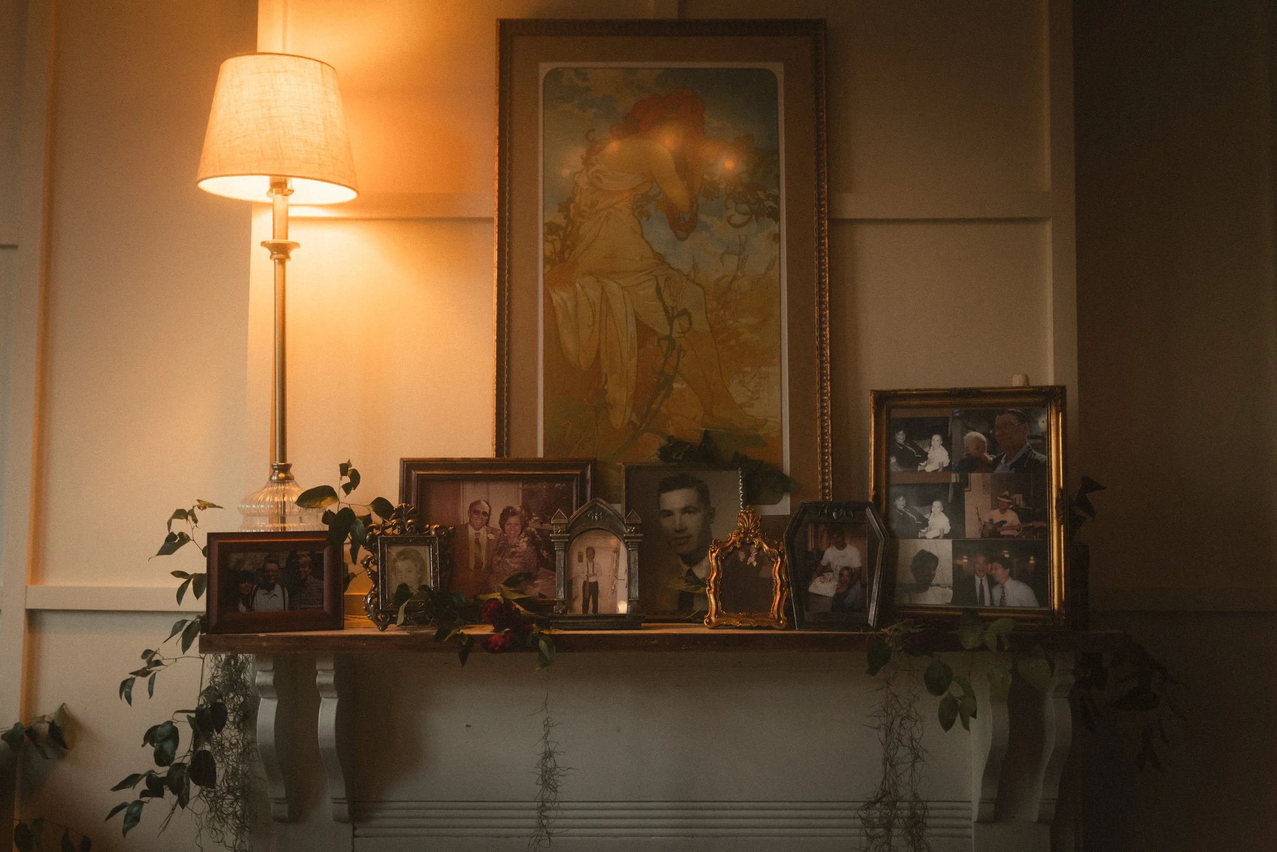 Warmly lit fireplace mantel decorated with framed family photographs, artwork, plants, and a table lamp