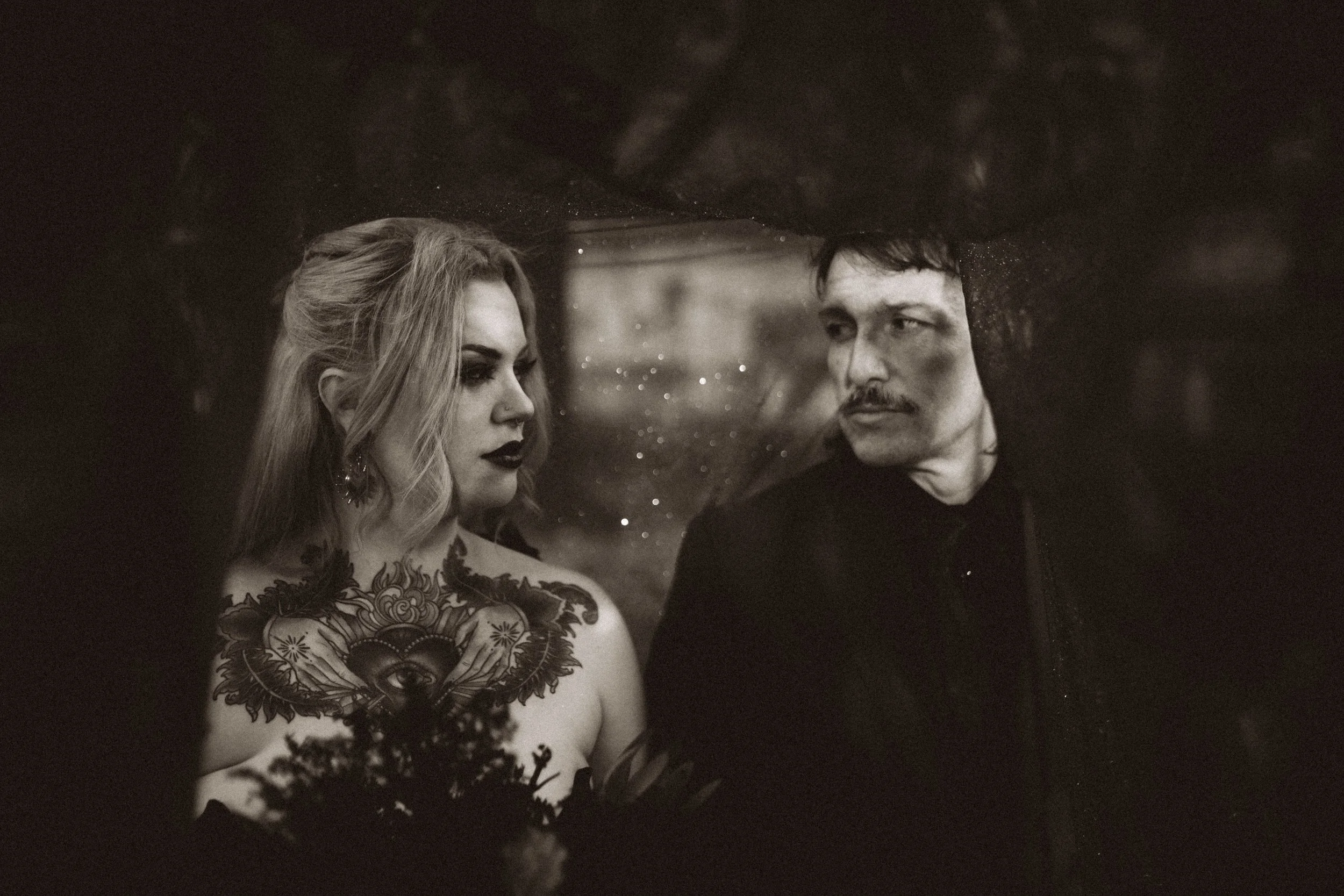 Black-and-white portrait of a tattooed woman and mustached man looking at each other through a rain-speckled window.