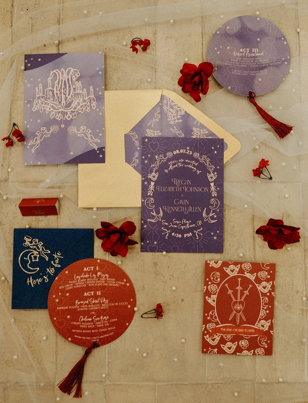 Wedding invitation display with multiple purple, red, and dark blue cards, decorated with white illustrations and text, surrounded by red flowers and small white pearls on a beige surface.