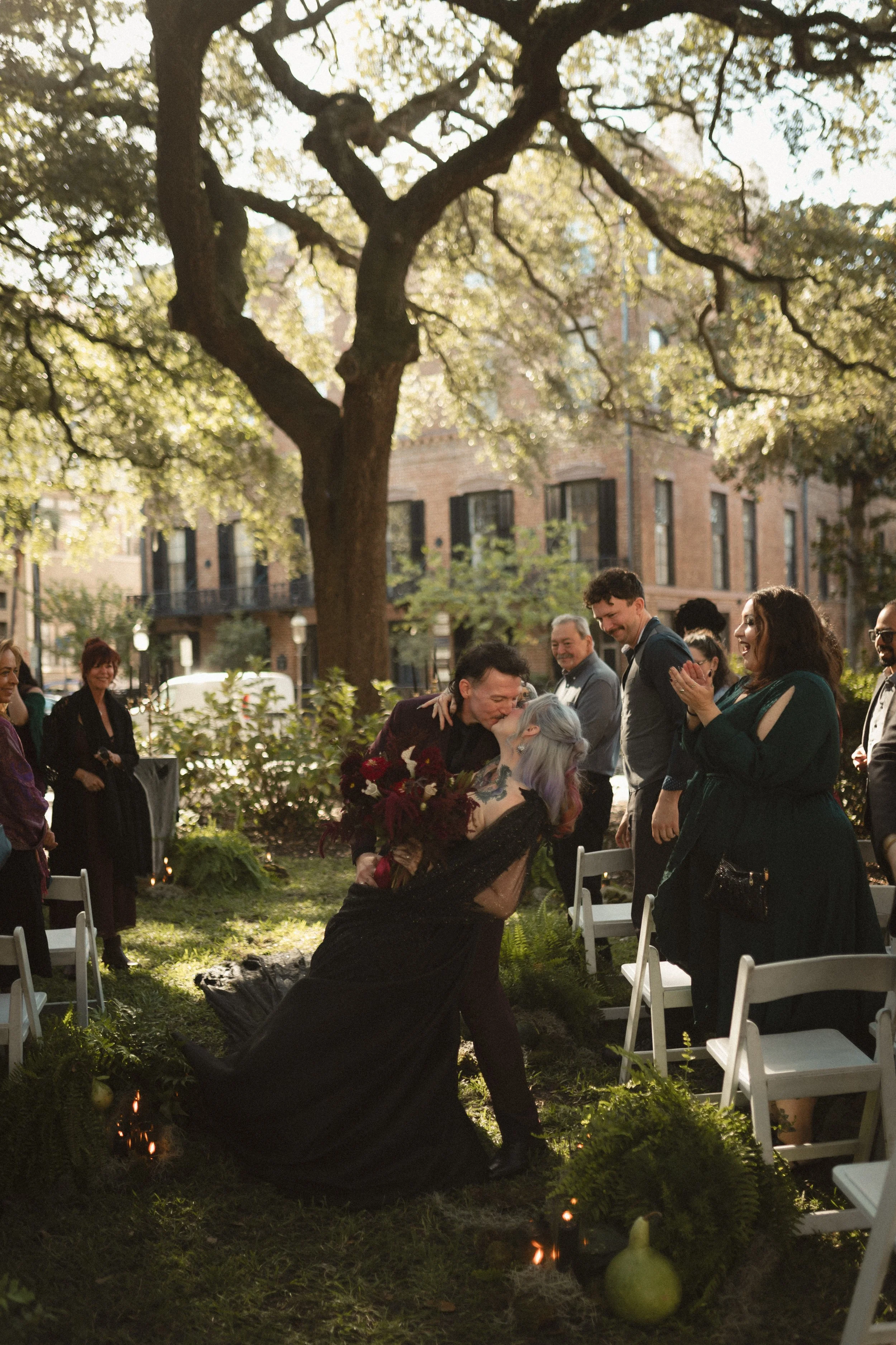 Outdoor wedding ceremony under large trees, with guests gathered around a couple embracing near white folding chairs.
