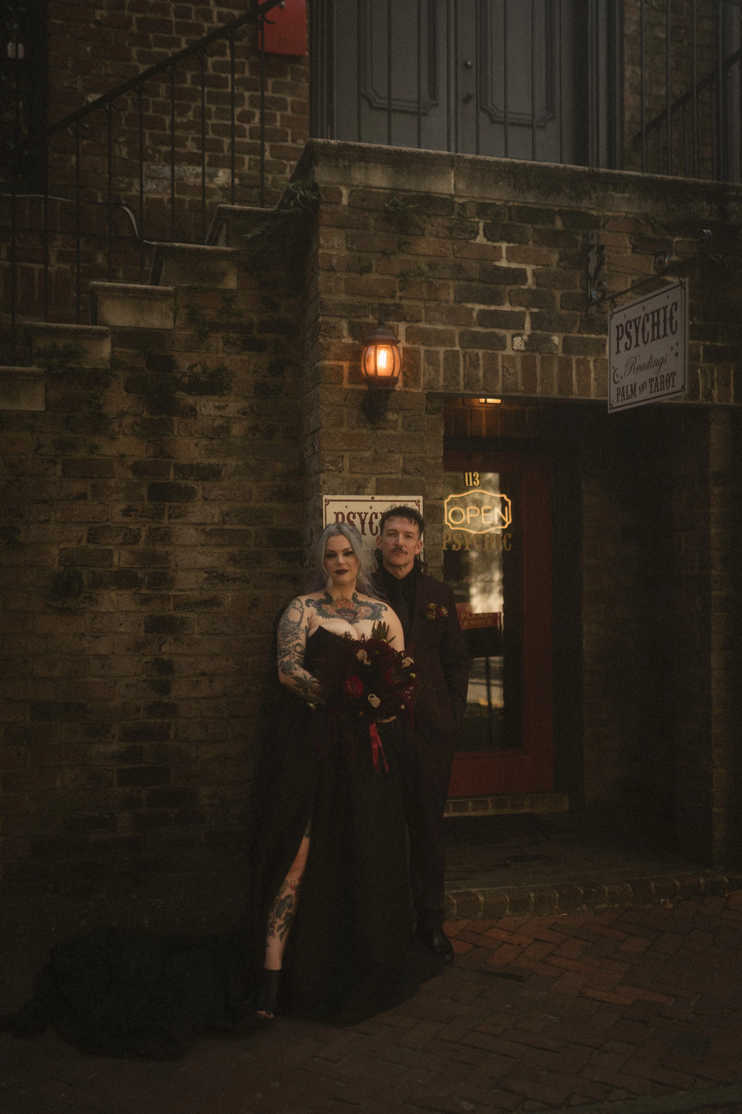 Tattooed woman holding a bouquet beside a man in black at a dimly lit brick-walled psychic reading venue.